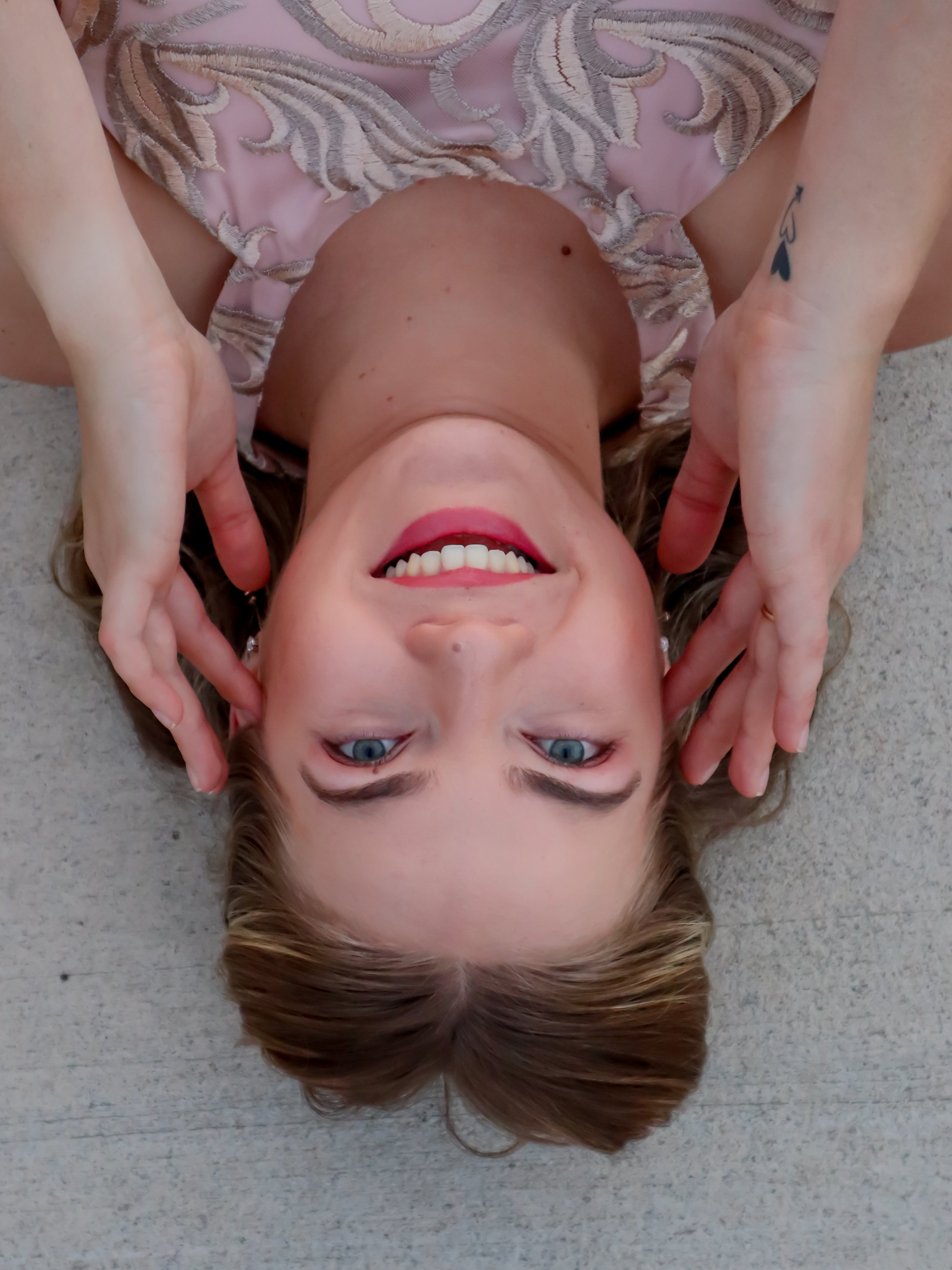 A woman lying on her back on the floor with her head upside down, smiling, with her hands framing her face, wearing a pink top with lace details.