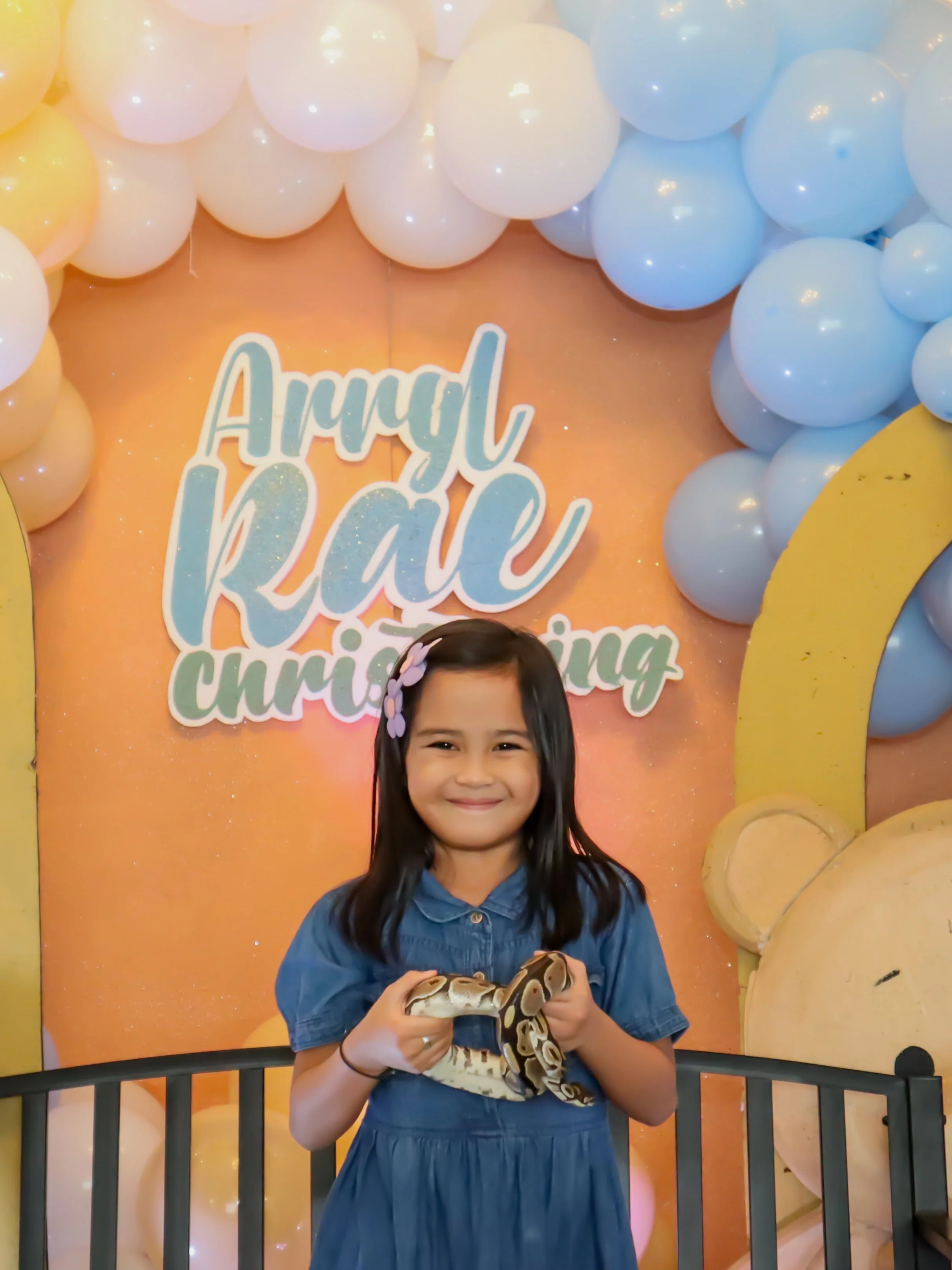 A young girl with long dark hair wearing a blue dress and pink butterfly hair clips, smiling while holding a snake in front of a colorful backdrop with balloons and a sign that reads 'April Rae celebrating'.