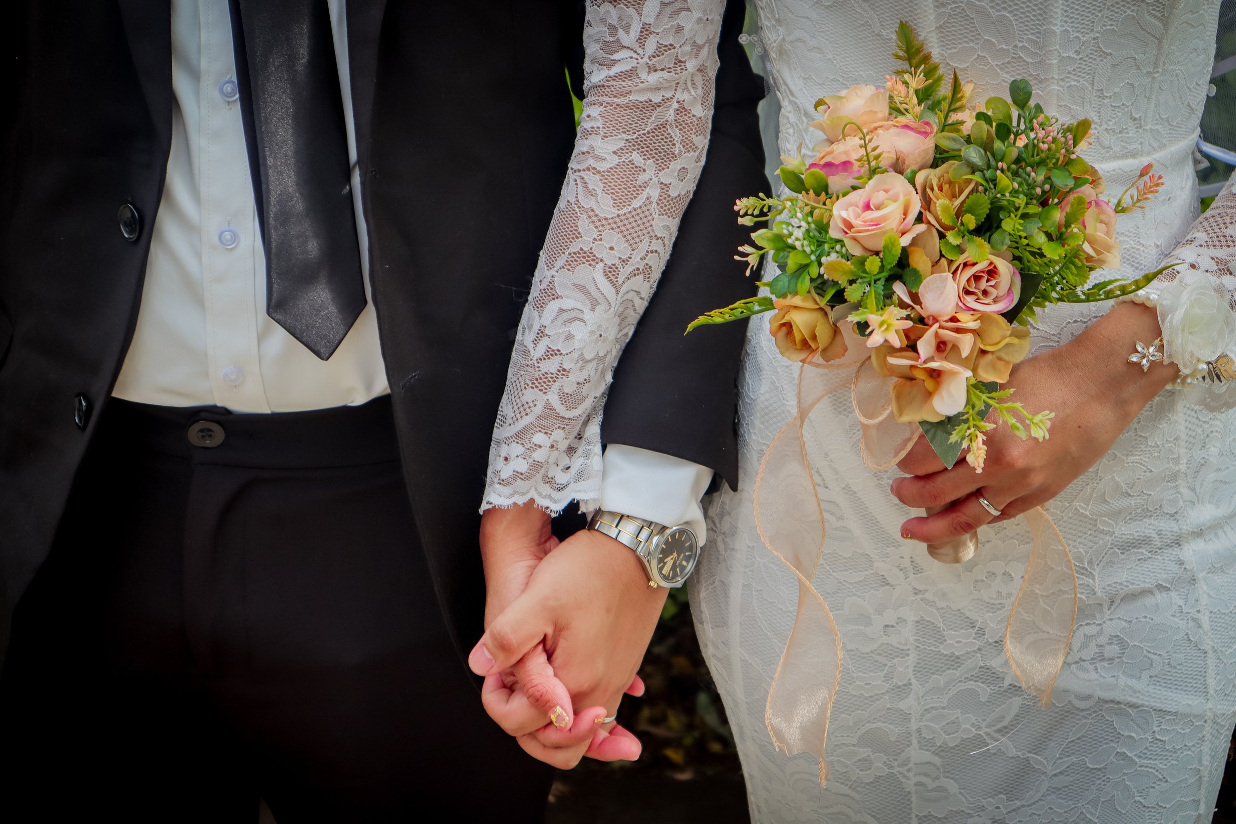 A bride and groom holding hands, with the bride holding a bouquet of peach roses and greenery, wearing wedding rings and a lace dress, and the groom in a dark suit with a watch.
