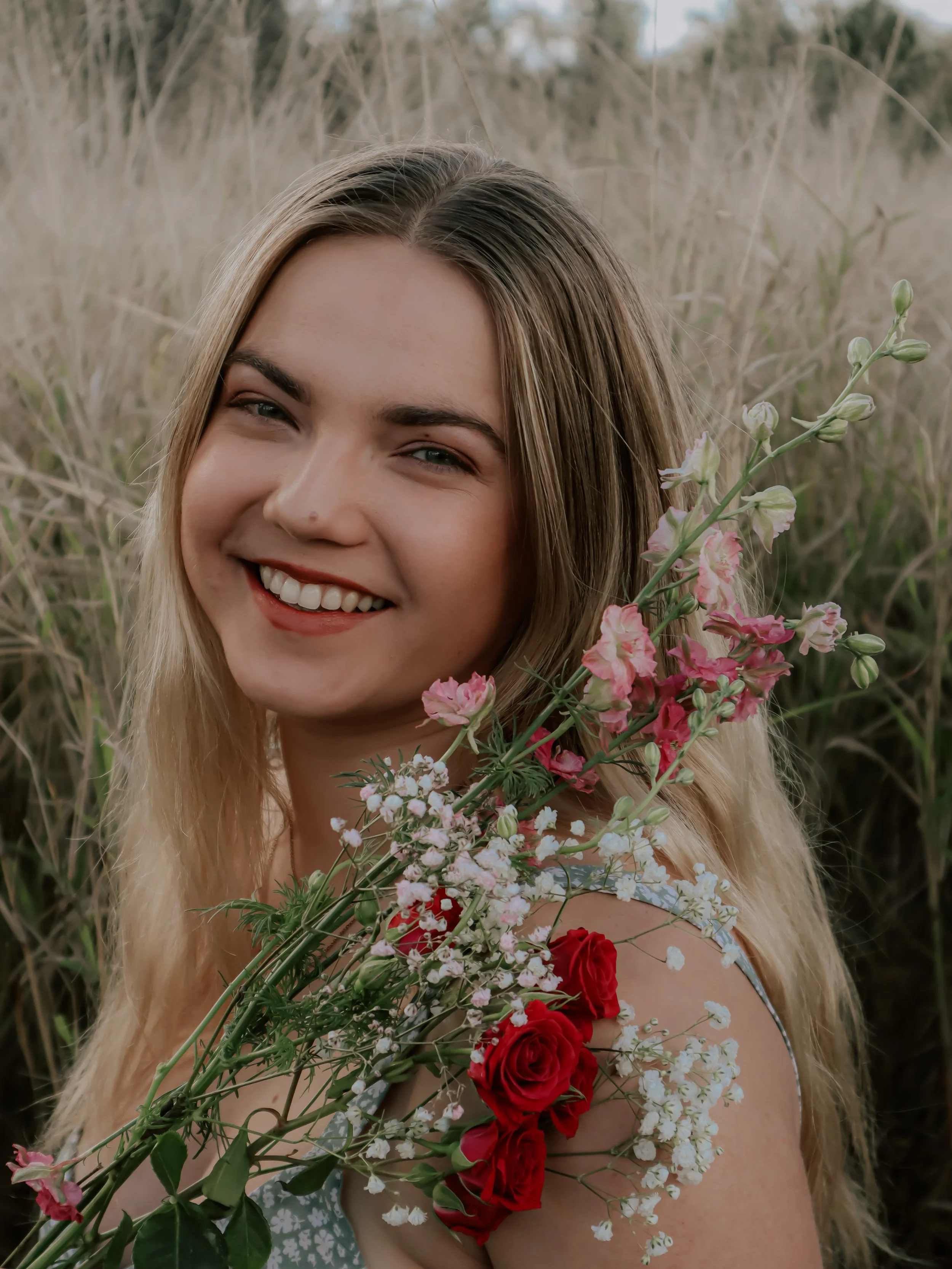 A young woman with long blonde hair holding a bouquet of pink and white flowers and wearing a floral dress, smiling in a field of tall grass.