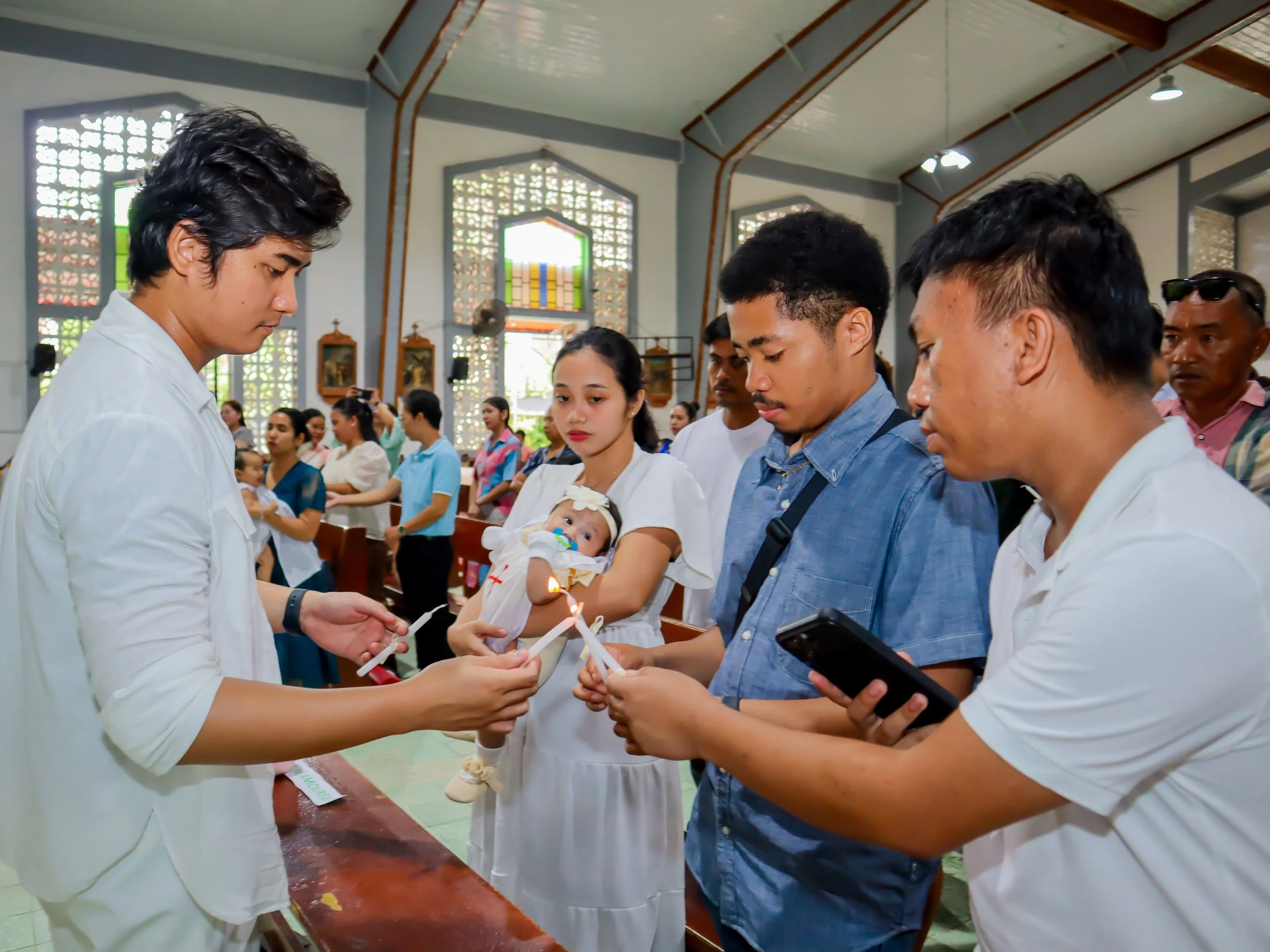 People participating in a baptism ceremony inside a church, lighting candles, with a woman holding a baby girl, and a group of individuals gathered around.