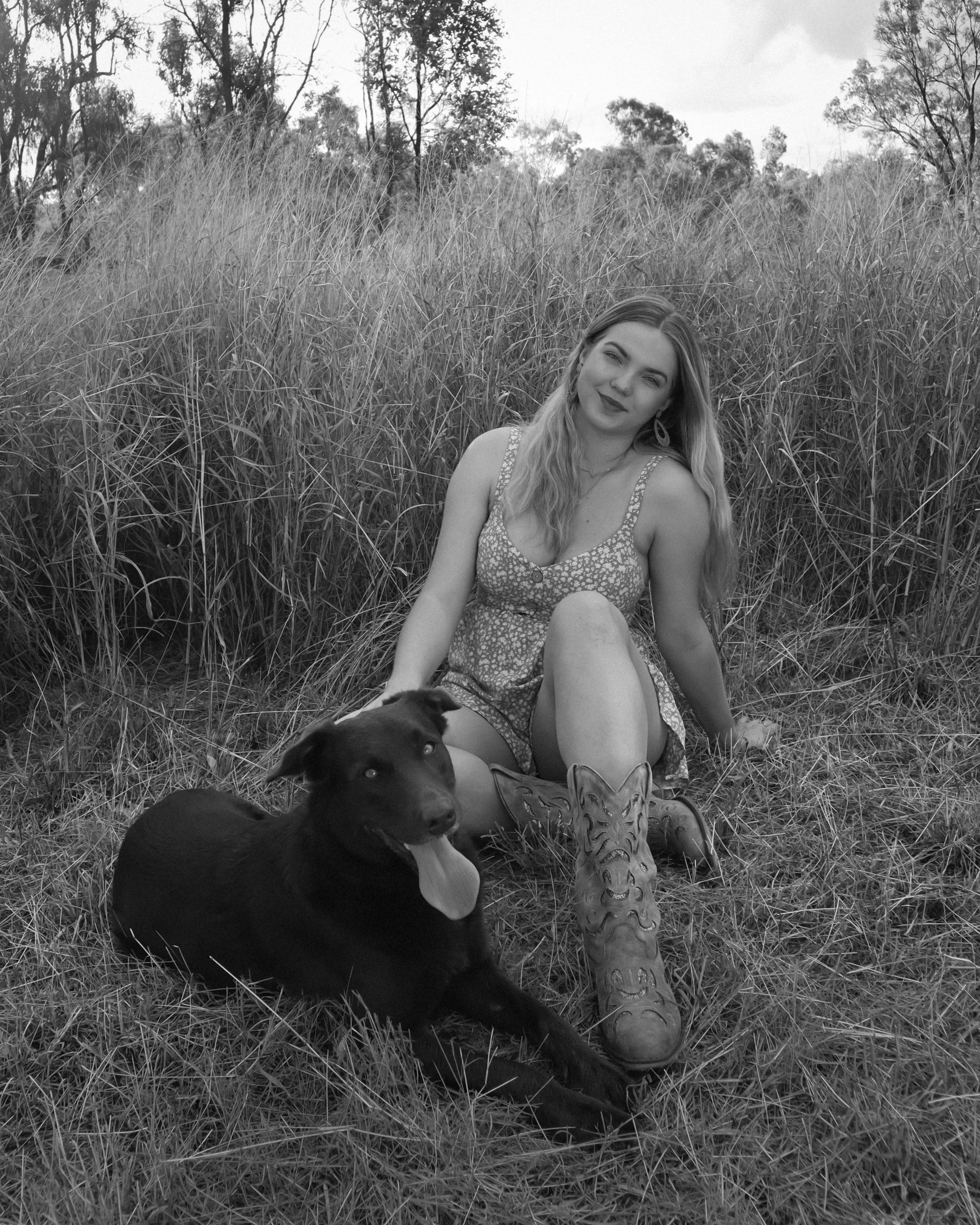 A woman with long hair sitting on the grass in a field with tall grass and trees in the background, posing with a black dog lying beside her, in black and white photograph.