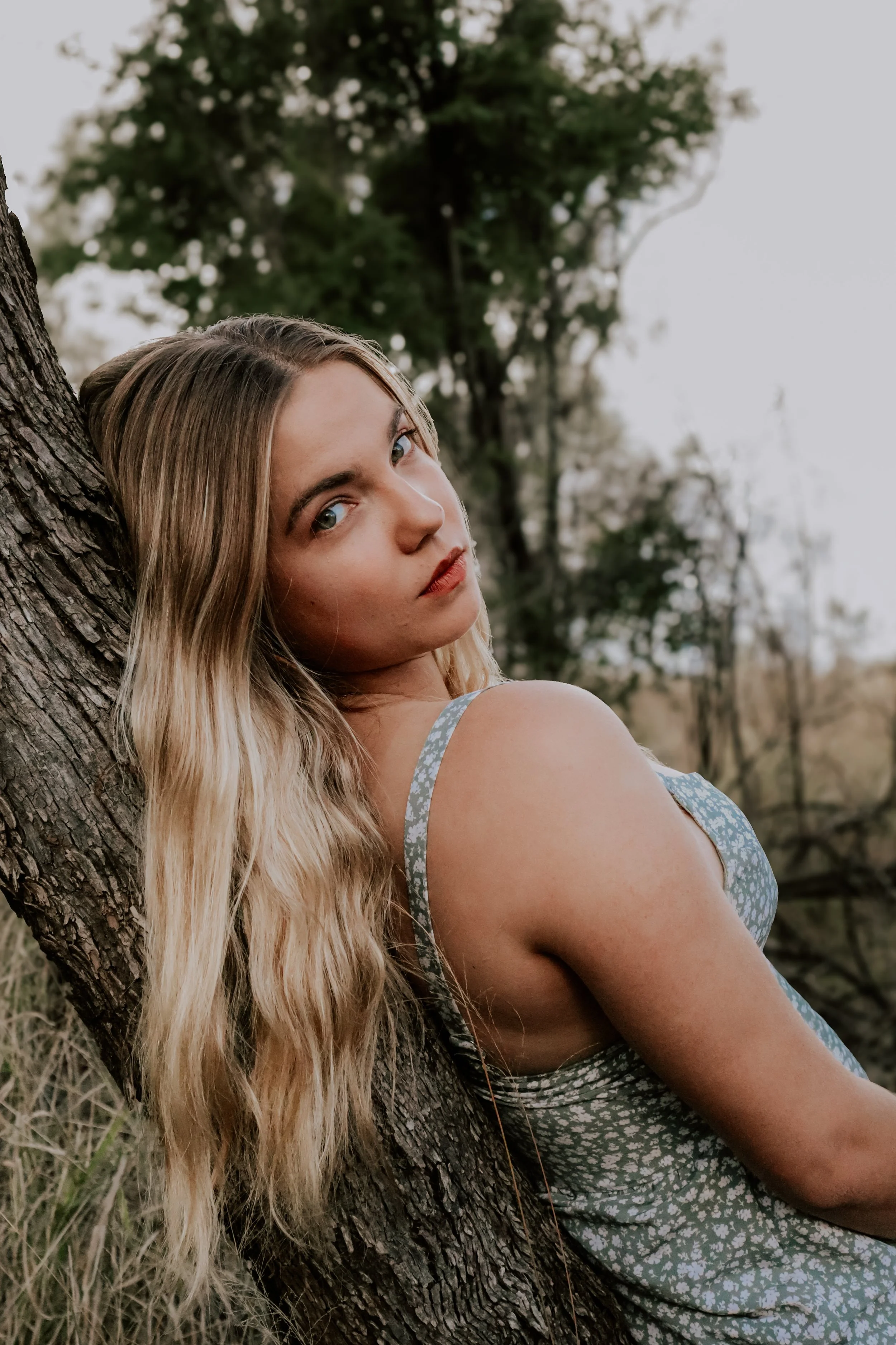 A young woman with long wavy blonde hair leaning against a tree outdoors, wearing a sleeveless dress with a floral pattern, looking at the camera.