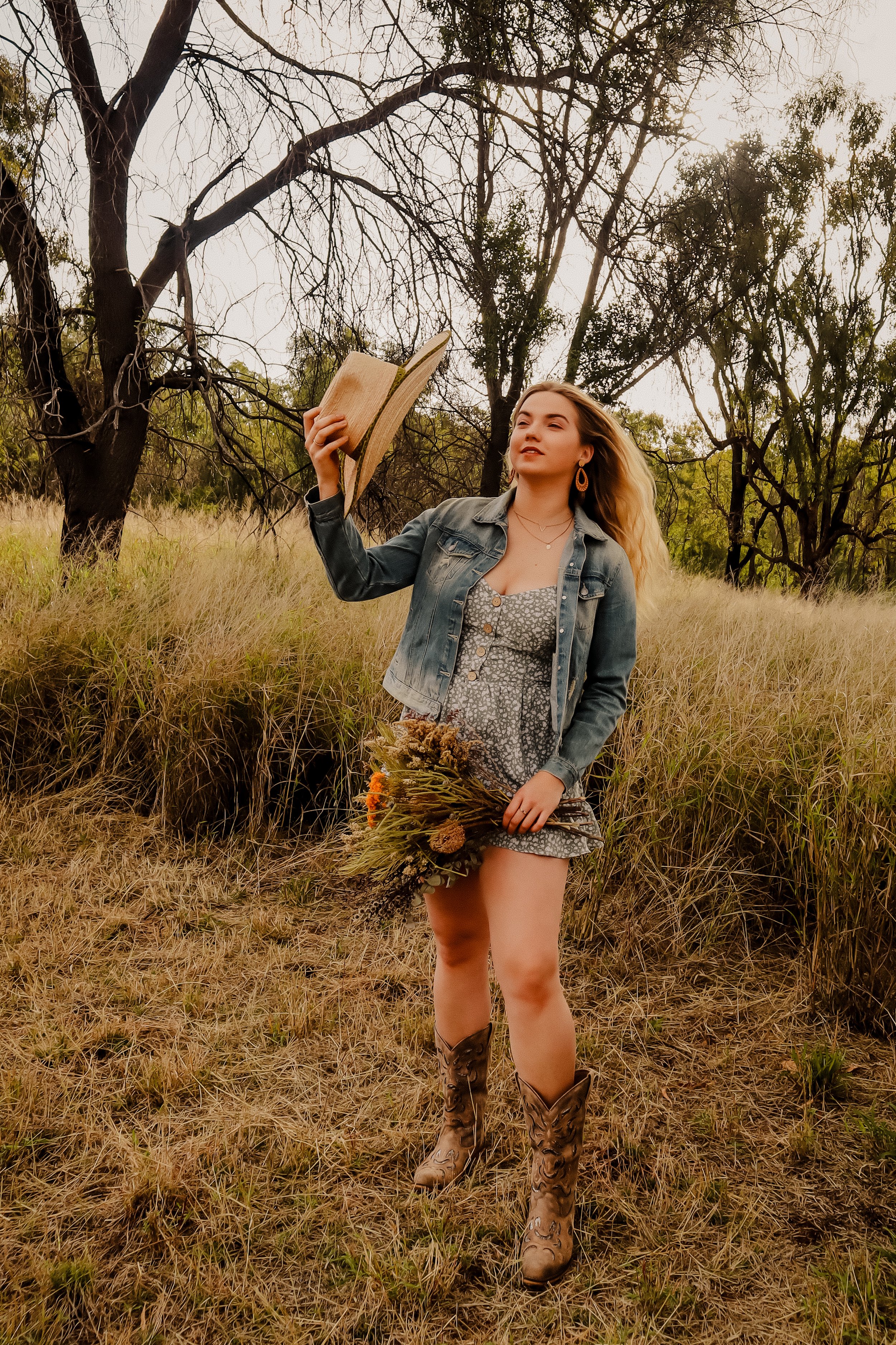 A young woman standing in a grassy field holding a bouquet of wildflowers in one hand and a straw hat in the other, wearing a grey floral dress, a denim jacket, and cowboy boots, with trees in the background.