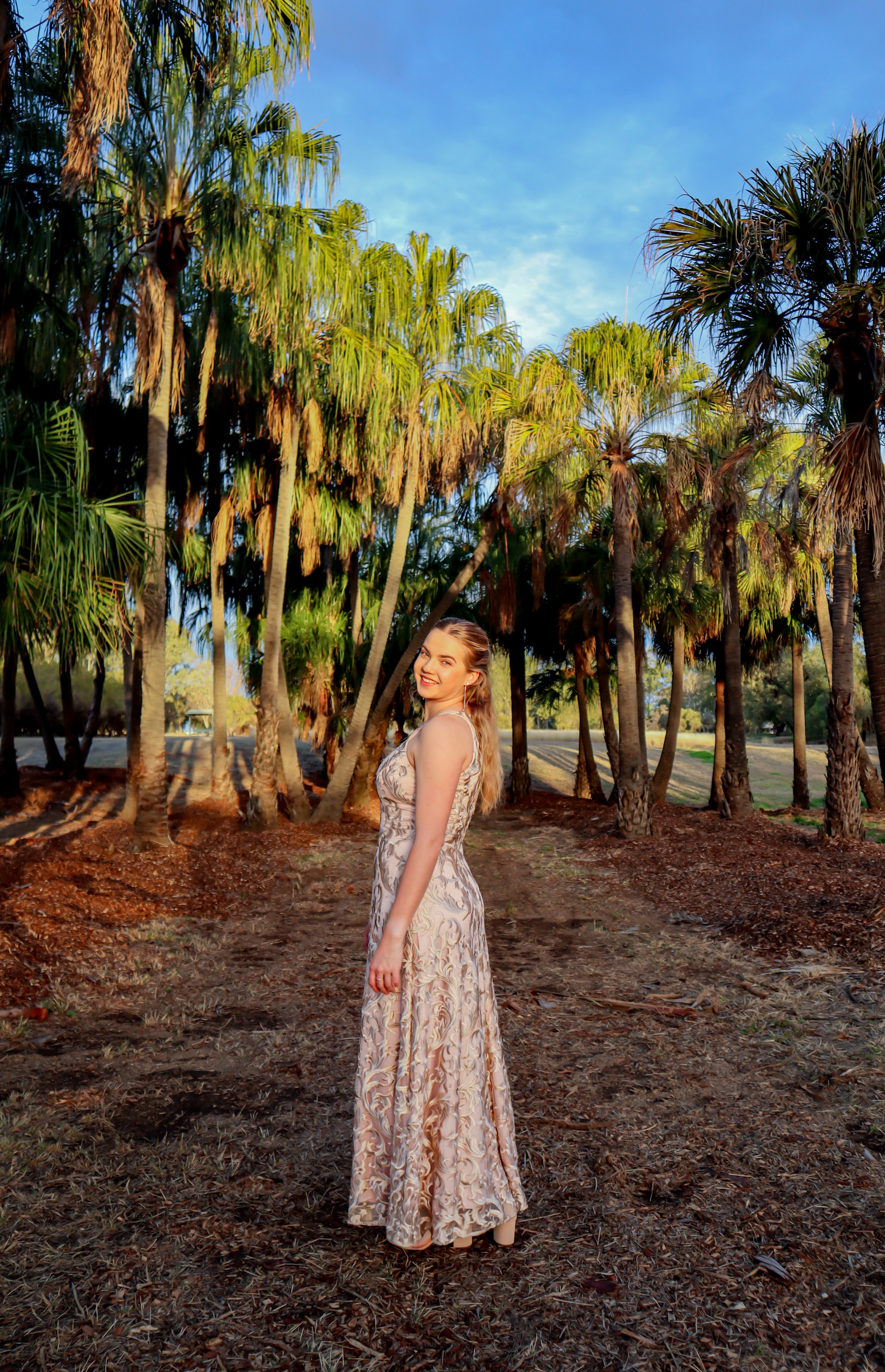 A woman in a long, elegant dress standing on a dirt path among tall palm trees under a partly cloudy sky, smiling at the camera.