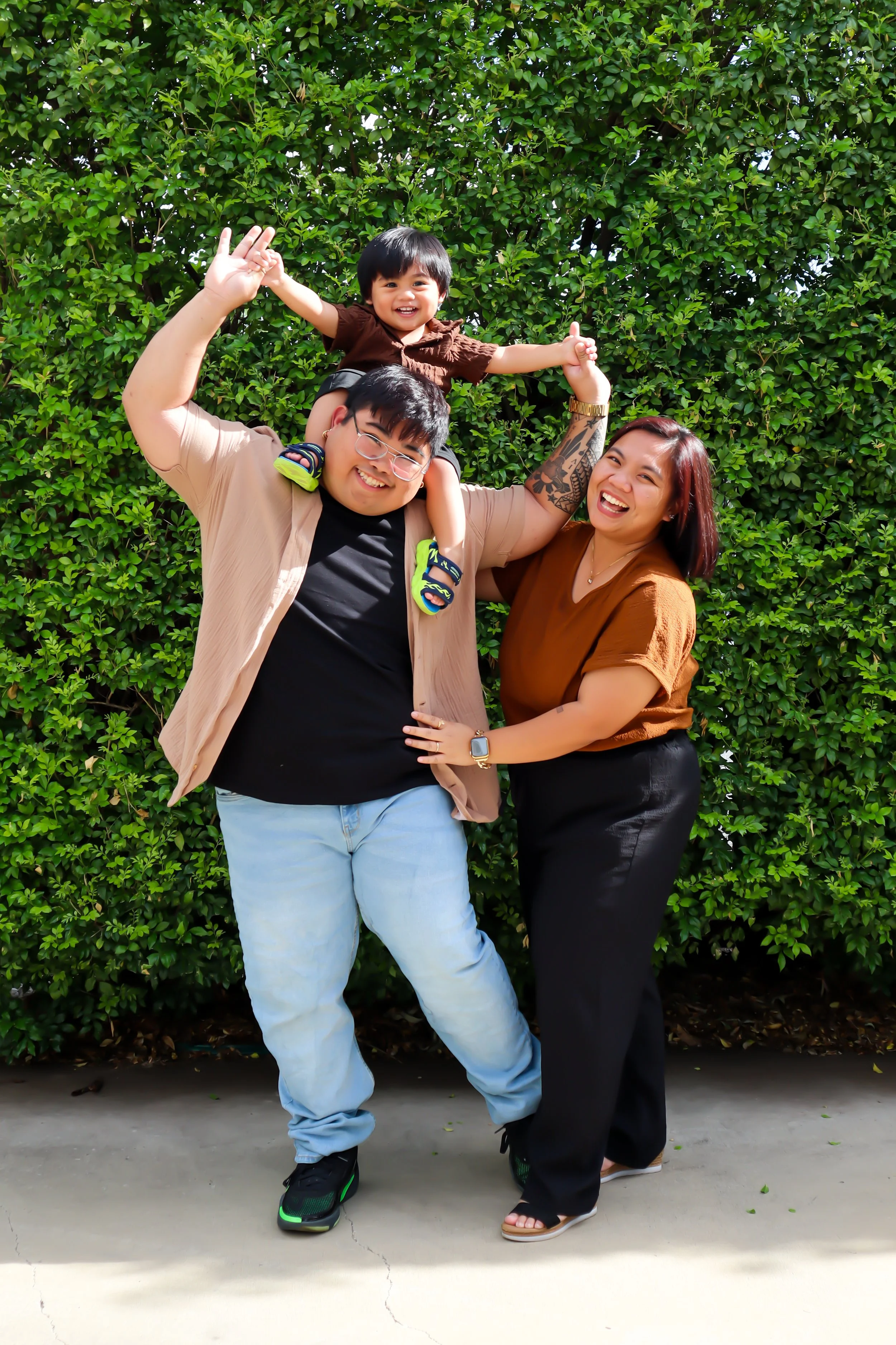 A joyful family group with a man, woman, and small child outdoors against a green leafy background. The man is holding the child on his shoulders, and the woman is standing beside them, all smiling and laughing.