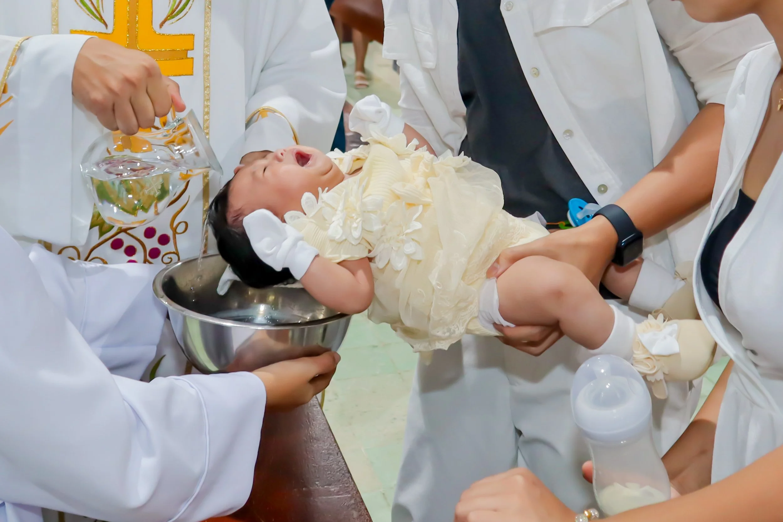 A baby being baptized, held by multiple people, with water poured over its head from a glass pitcher, in a ceremonial religious setting.