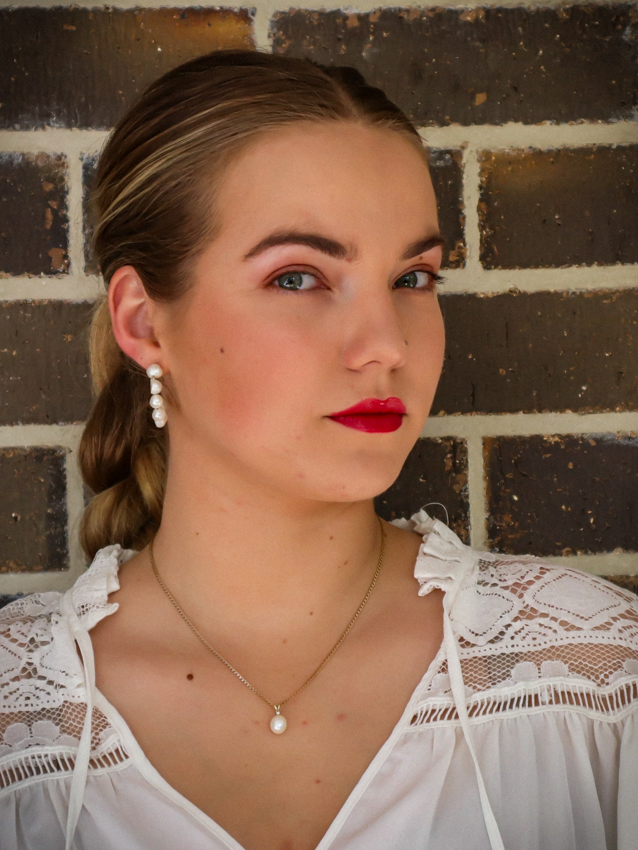 A young woman with blonde hair tied back, wearing pearl earrings, a pearl necklace, and a white blouse with lace details. She has red lipstick and is standing against a brick wall.