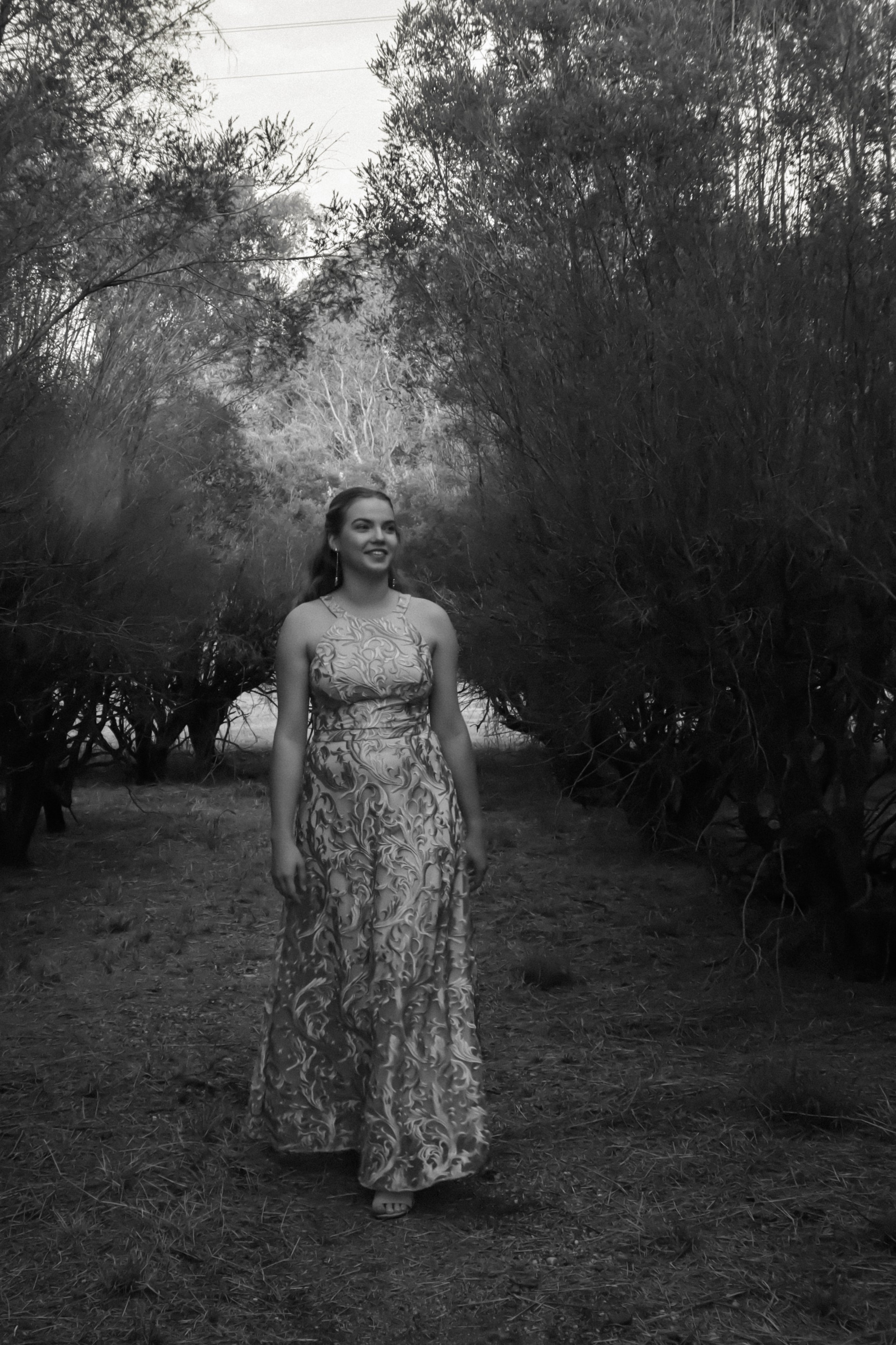 A young woman in a patterned dress walking through a wooded area, black and white photograph.