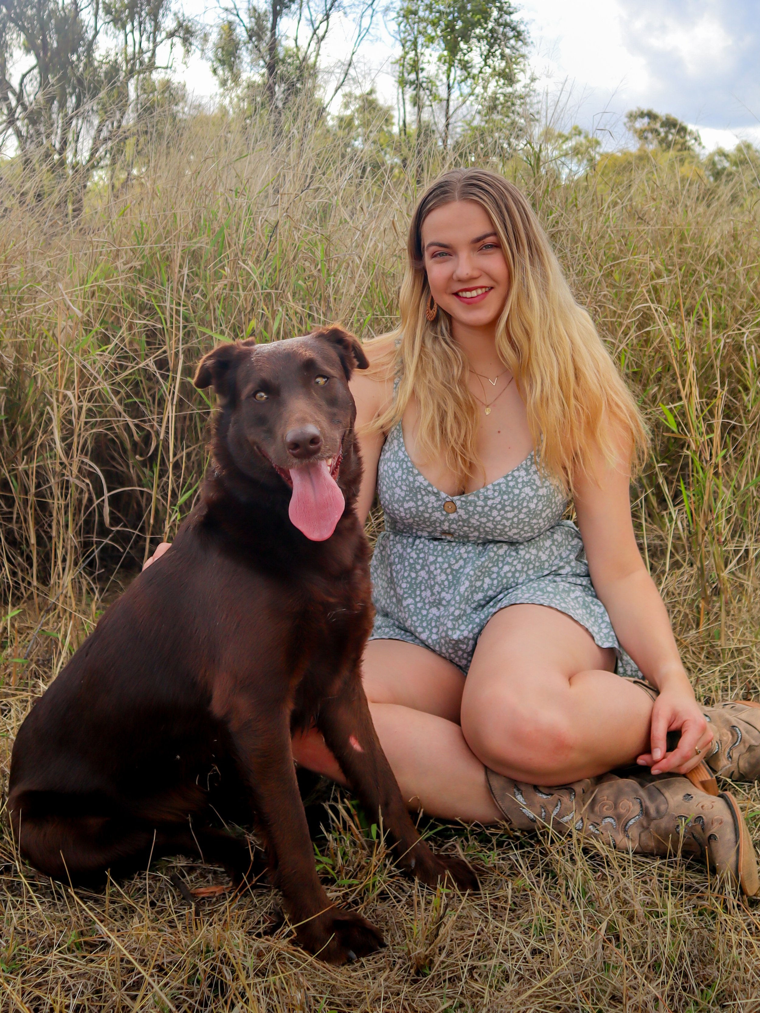 A young woman with long blonde hair, wearing a gray dress with a floral pattern, is sitting on the grass next to a brown dog with its tongue out. They are outdoors in a field of tall grass with trees in the background.