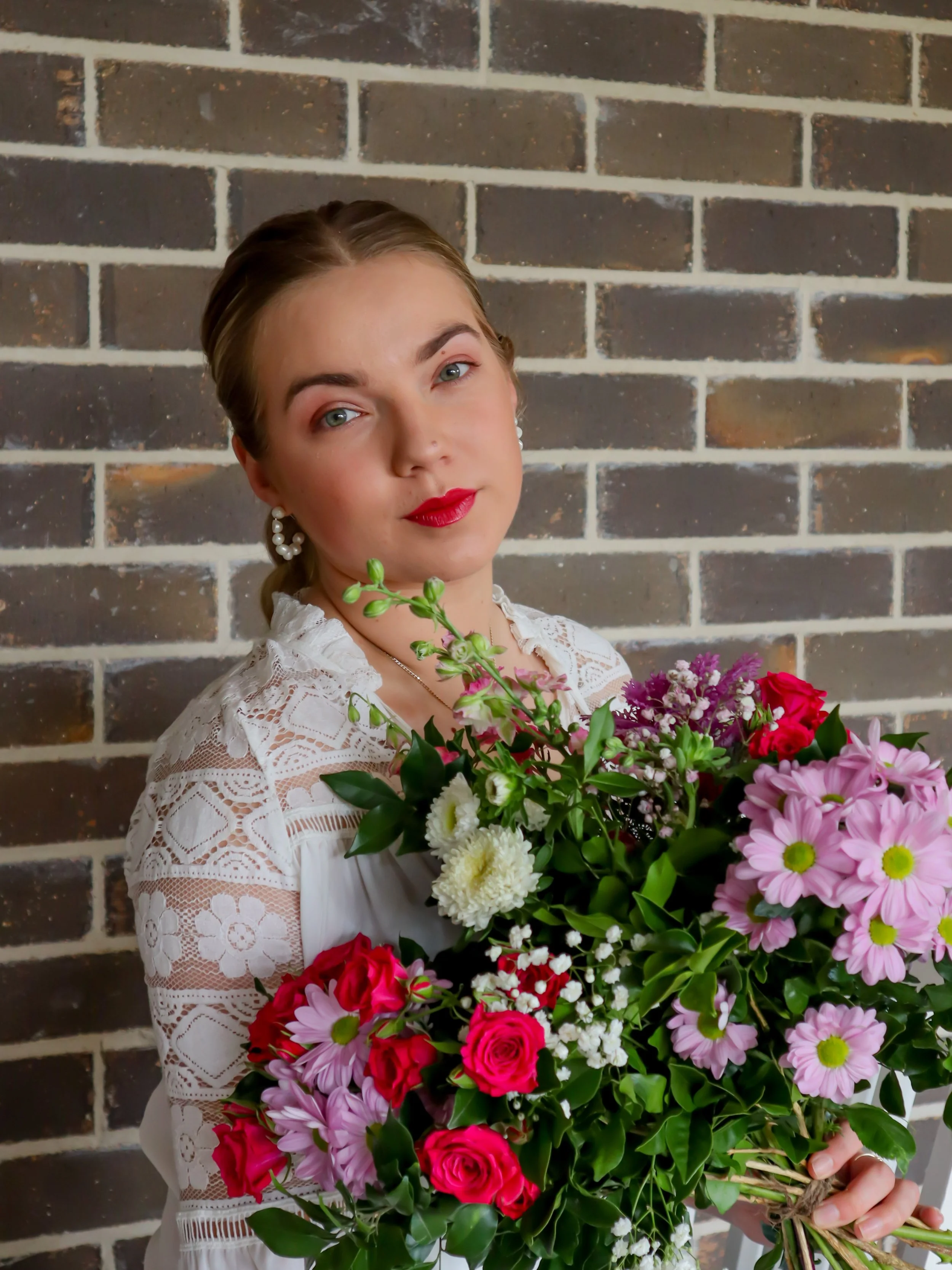A woman with blonde hair and red lipstick holding a large bouquet of pink, red, white, and purple flowers in front of a brick wall.