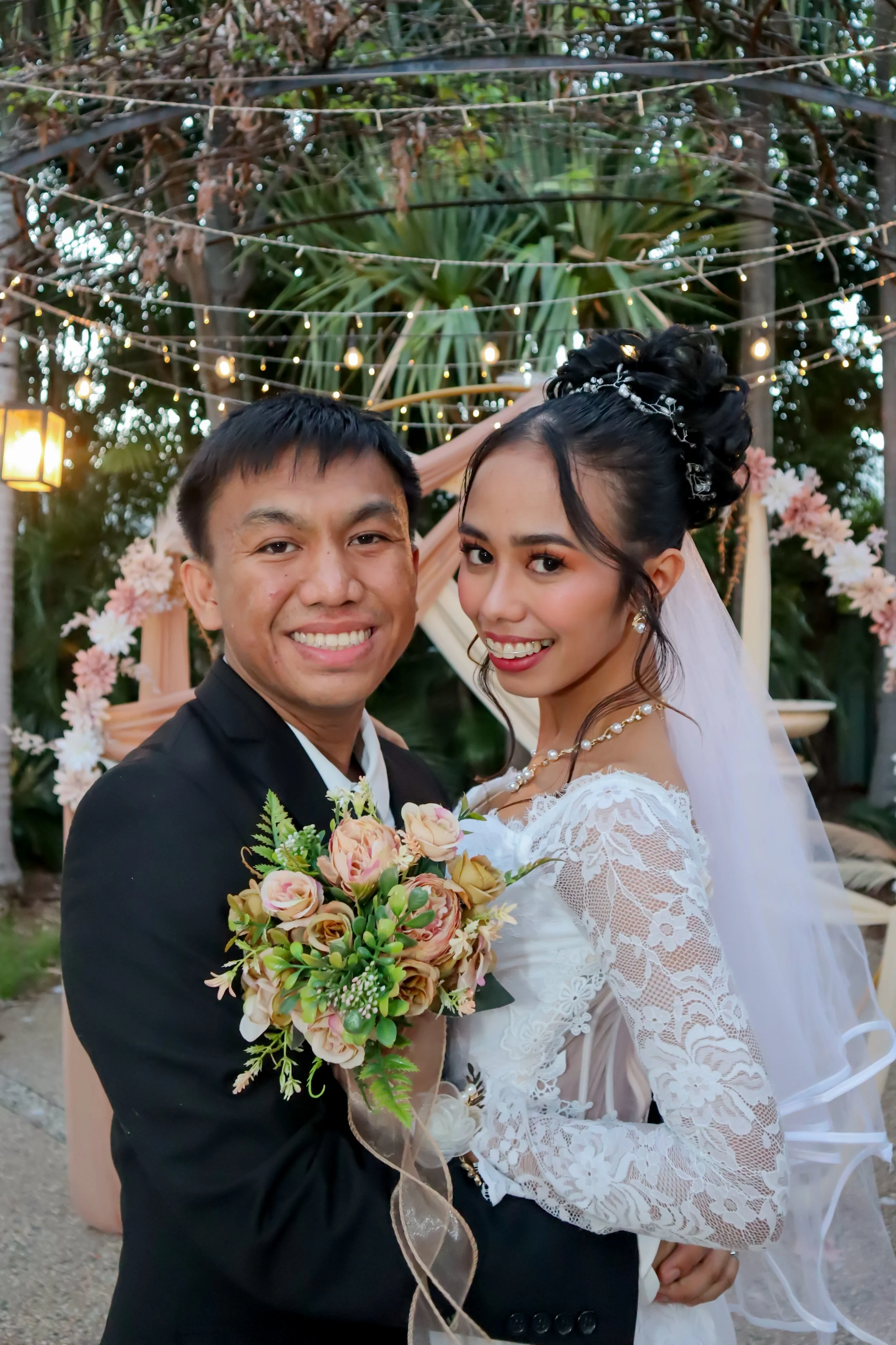 A wedding couple smiling outdoors, woman in a white lace wedding gown with veil and pearl jewelry holding a bouquet, man in black suit, decorated with string lights and floral arrangements in the background.