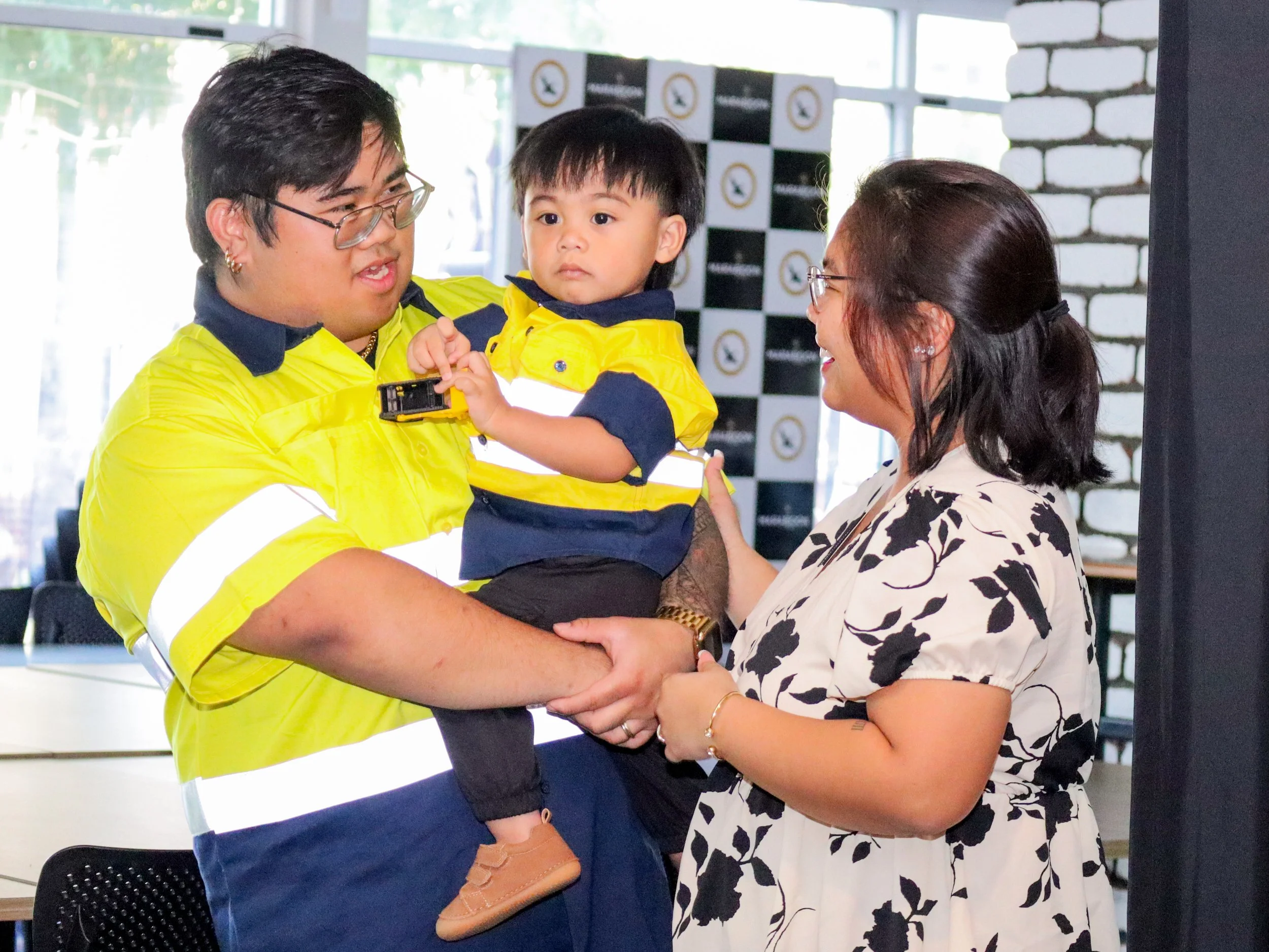A man in a yellow high-visibility jacket holding a young boy, and a woman smiling and reaching out towards them, in an indoor setting with large windows and white brick walls.