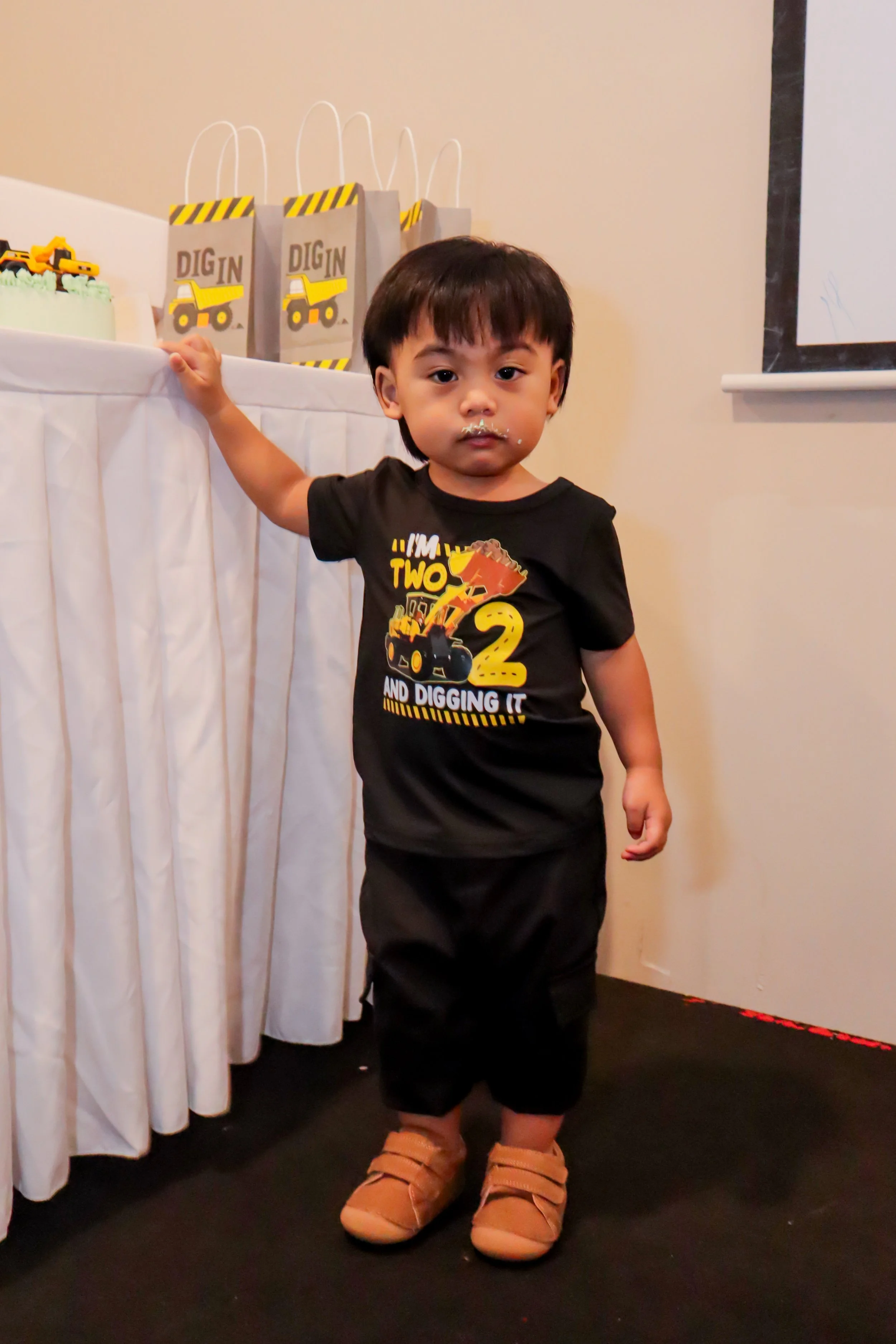 A young child with dark hair and brown skin, wearing a black T-shirt with construction artwork and the number 2, and tan shoes, stands indoors near a cake and party favor bags.