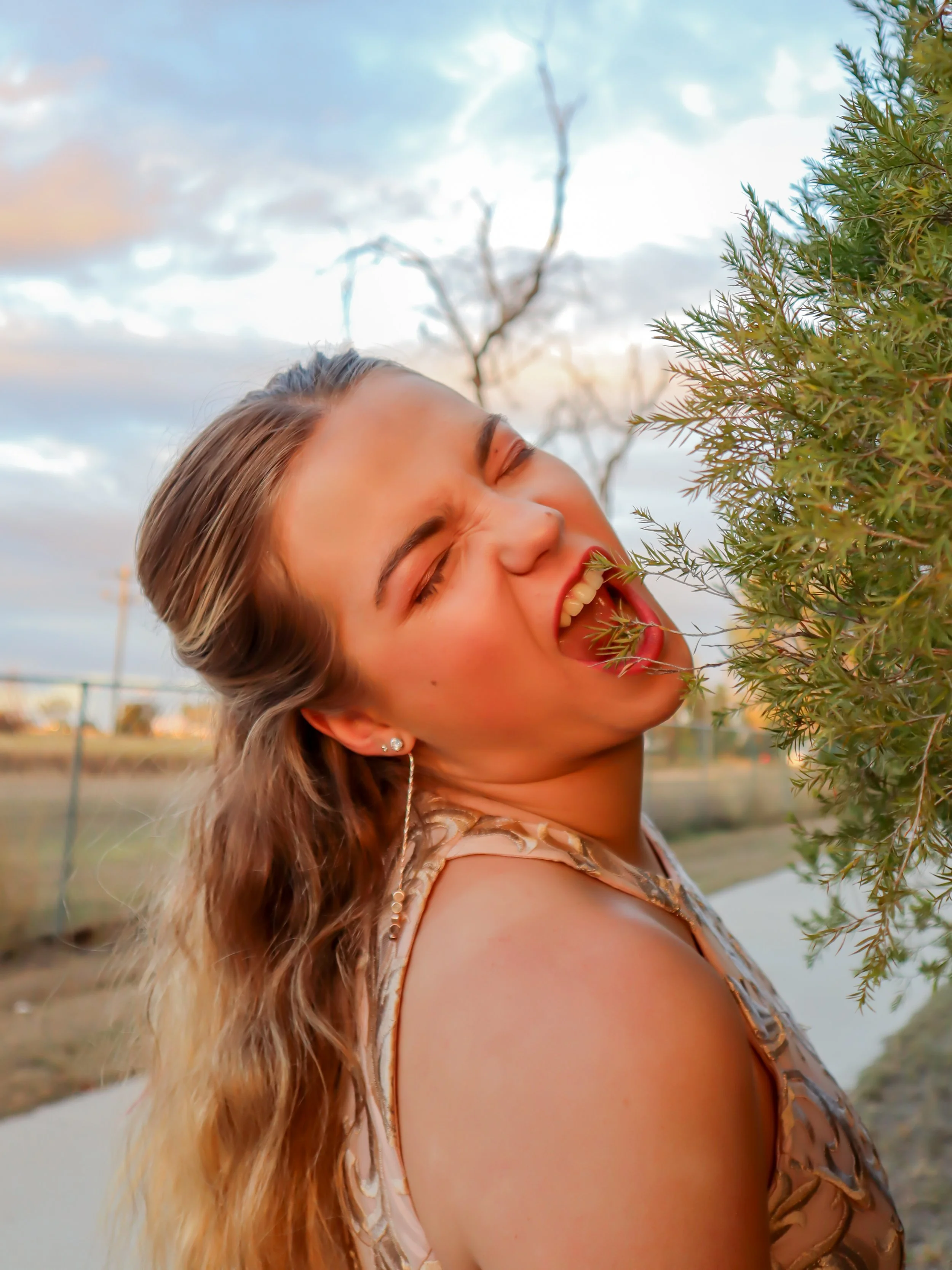 A young woman with long, wavy hair is outdoors, smiling with her eyes closed and sticking her tongue out as she touches a bush with a branch. The sky is partly cloudy with some trees in the background.
