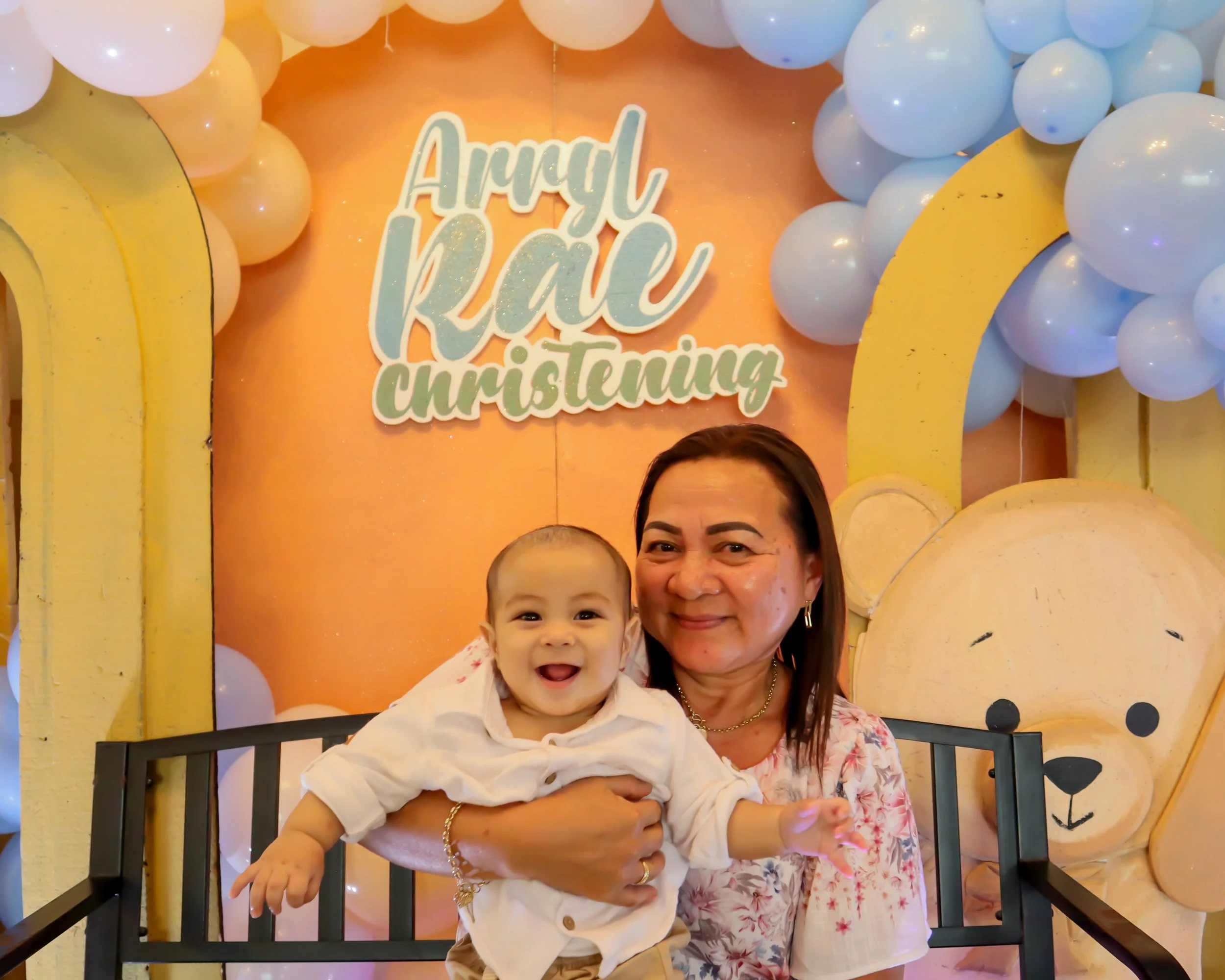 A woman holding a smiling baby in front of a birthday party backdrop decorated with balloons, a sign that reads 'Arugh Kai Christmas', and a large teddy bear illustration.