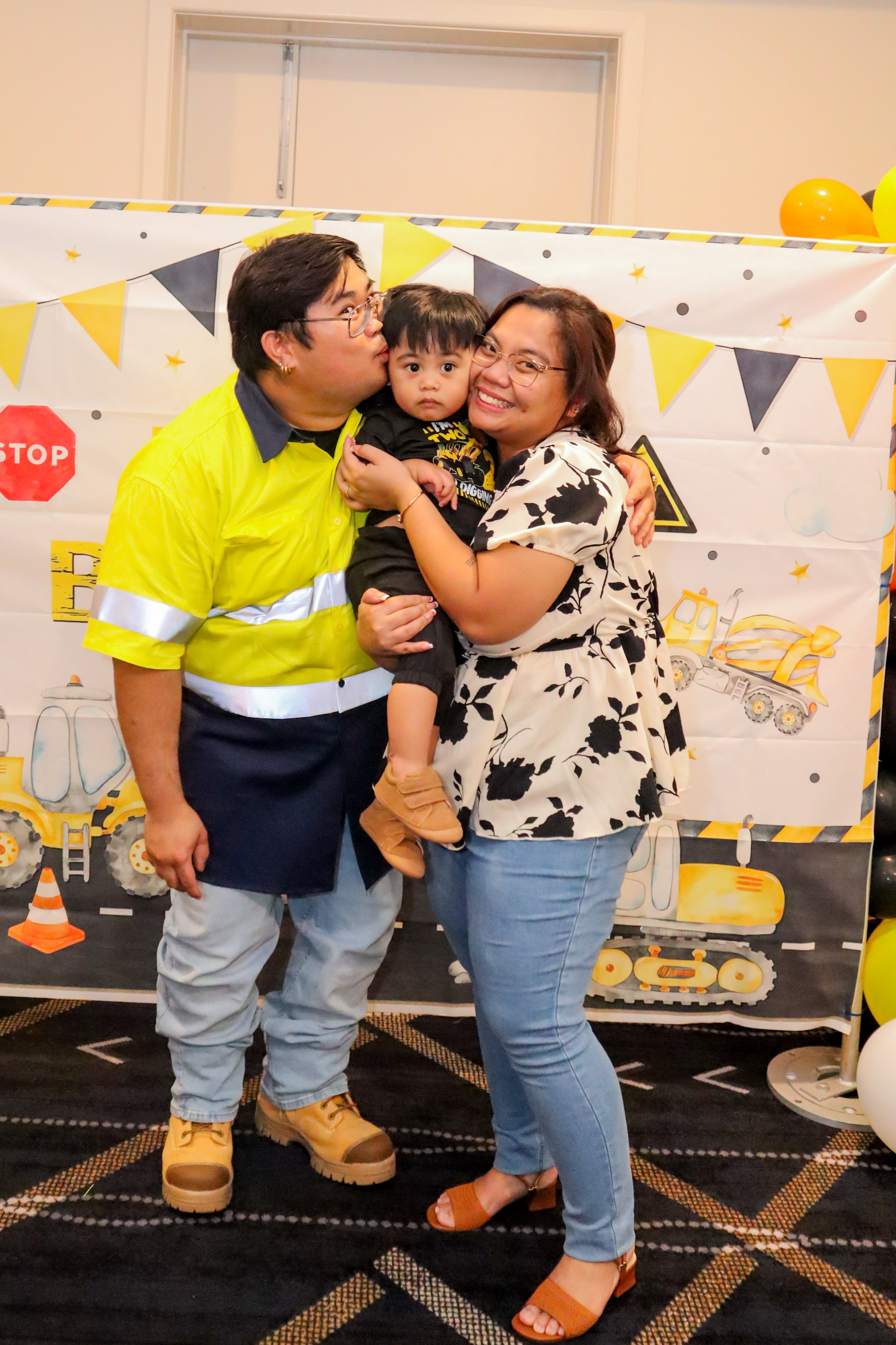 A family of three hugging in front of a construction-themed backdrop with yellow and black balloons.