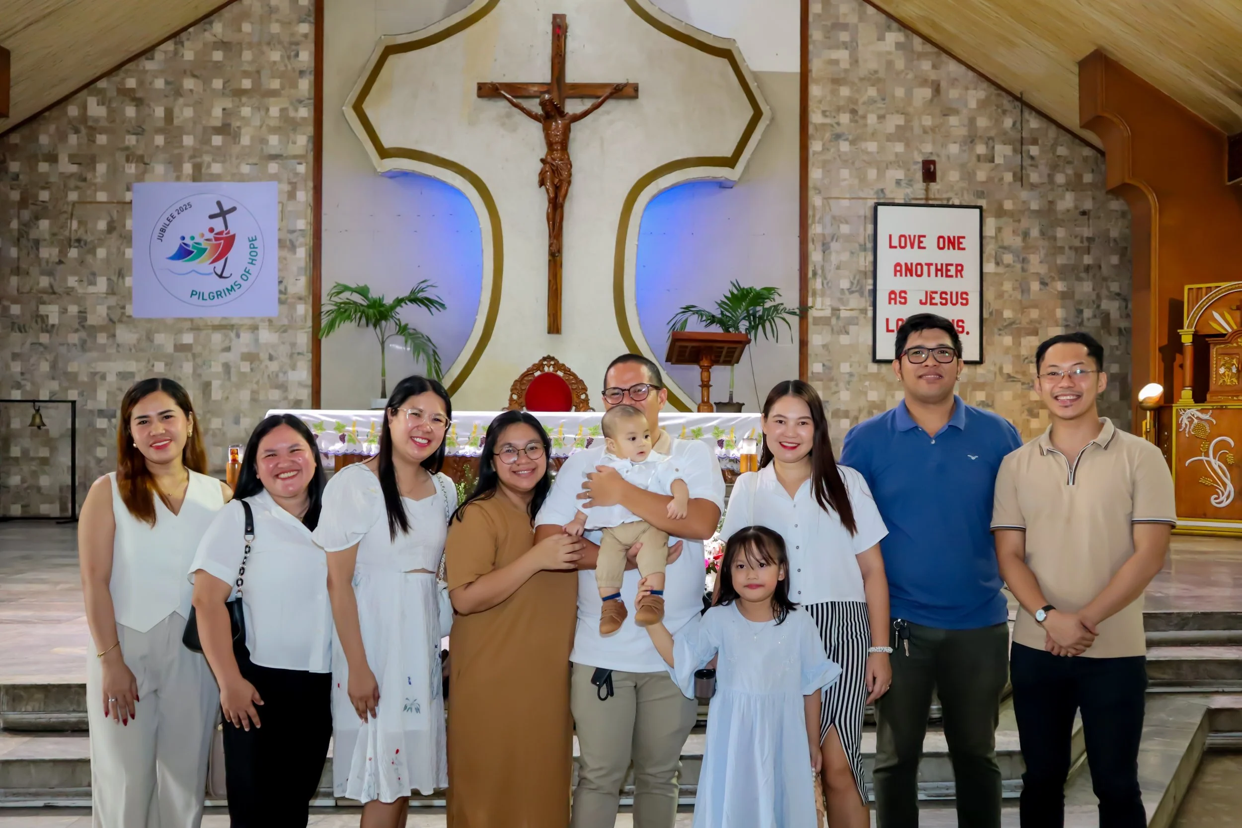 Group of people, including children and adults, gathered in a church for a religious ceremony, standing in front of a large crucifix with a gold outline on the wall.