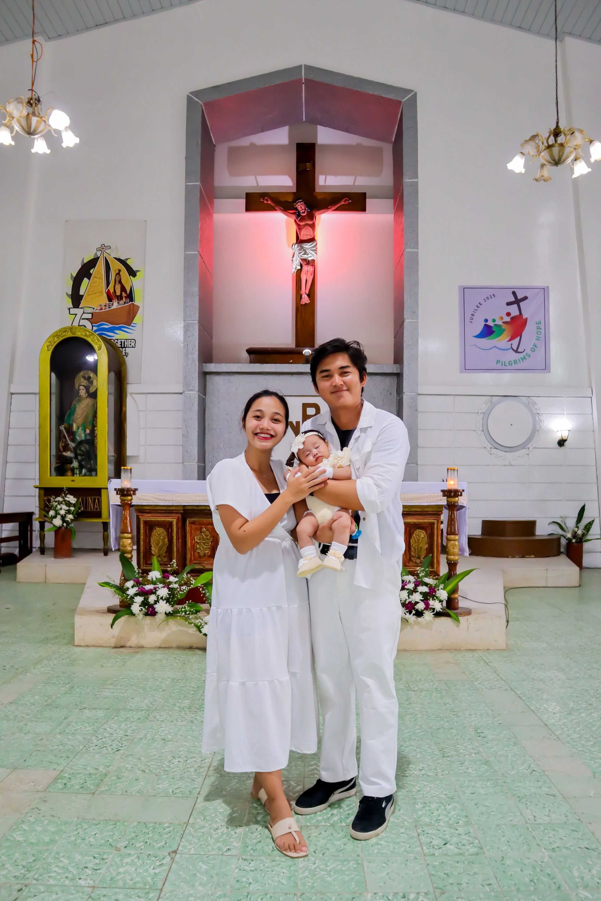 A family of three standing inside a church, with the mother, father, and their baby during a baptism ceremony. The mother and father are smiling and holding their baby, all dressed in white. The church has a crucifix on the altar, floral arrangements