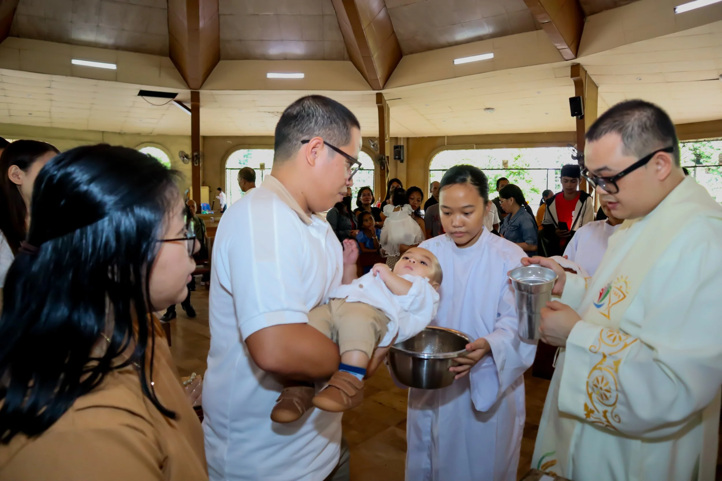A baby being baptized in a church, surrounded by family members and a priest holding a small bowl.