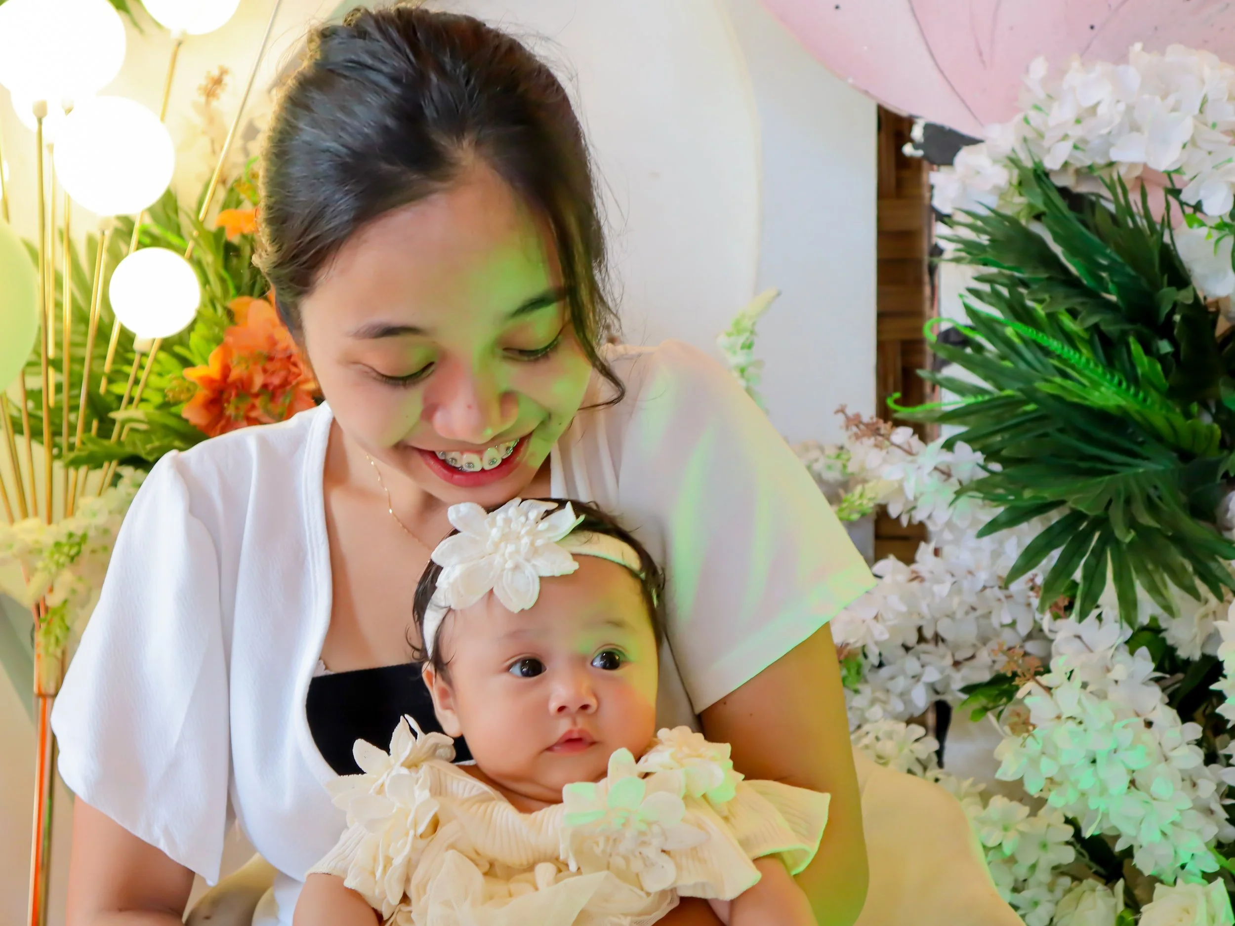 A young woman with dark hair smiling while holding a baby girl, surrounded by white flowers and green foliage, at a celebration or party.
