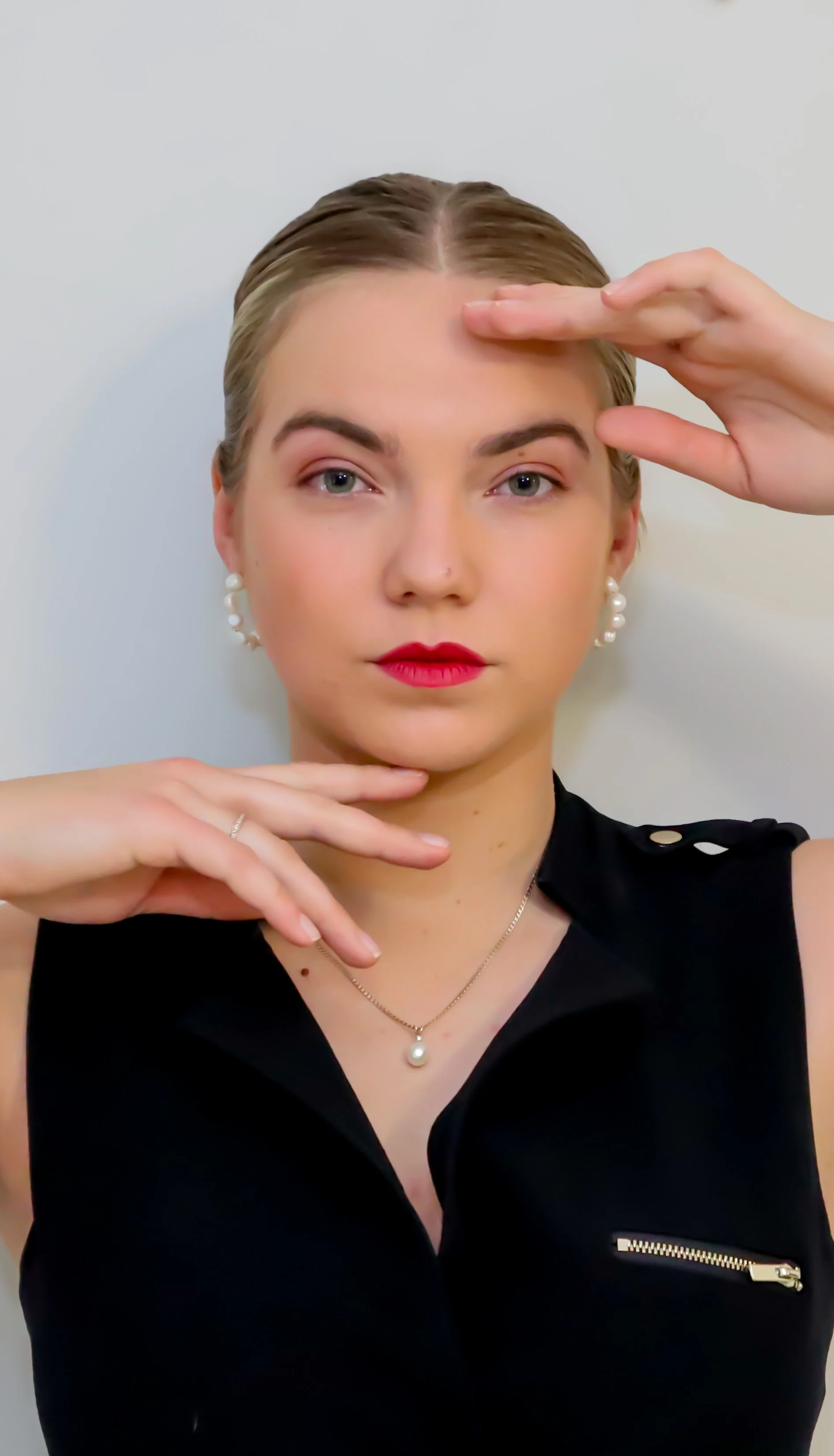 A woman with blonde hair styled back, wearing pearl earrings and a pearl necklace, dressed in a black sleeveless top with gold accents, posing with her hand on her forehead and chin against a plain light background.