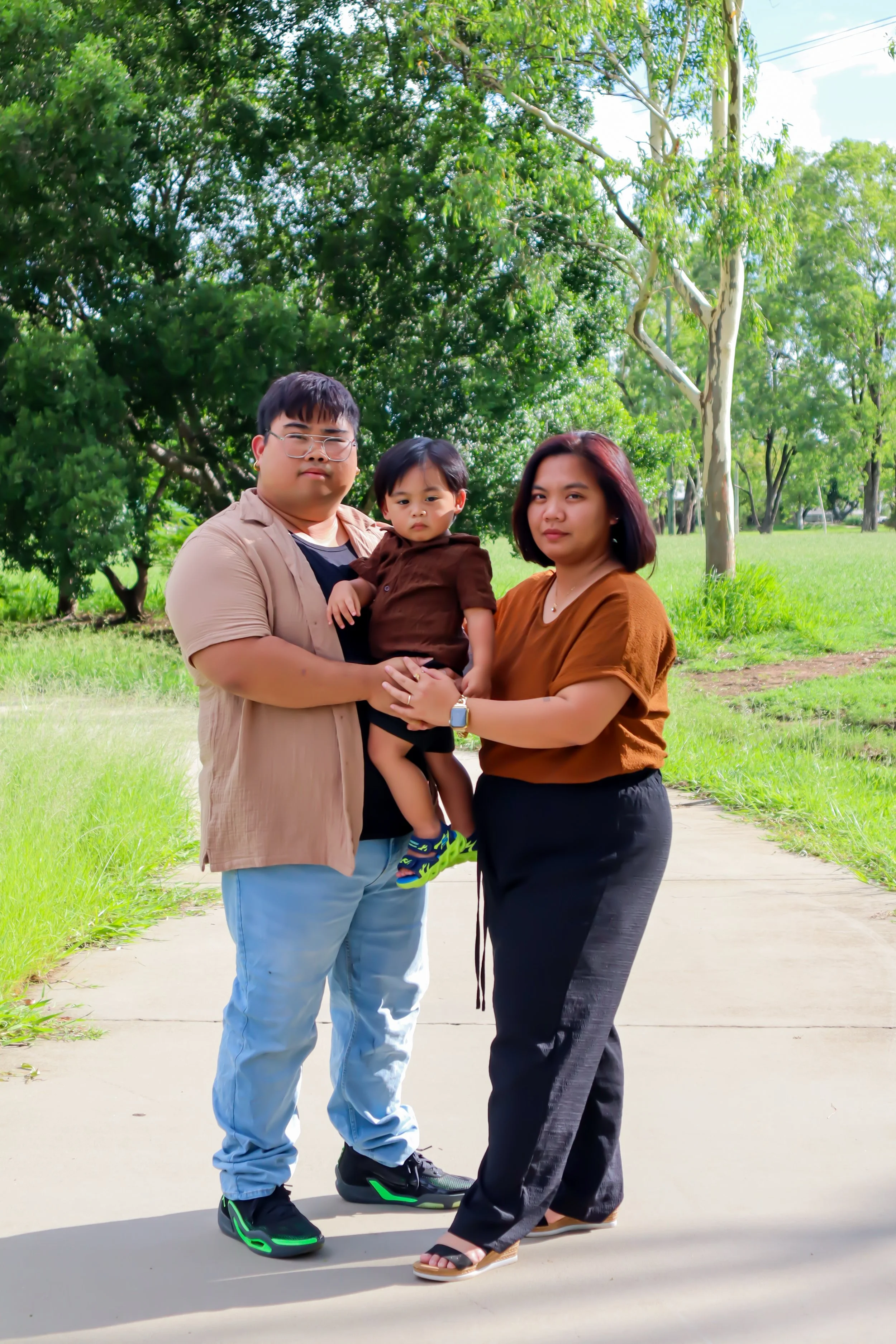 A family of three standing on a park pathway with green grass and tall trees in the background. The father is holding a young child, and the mother is standing beside them, all looking at the camera.