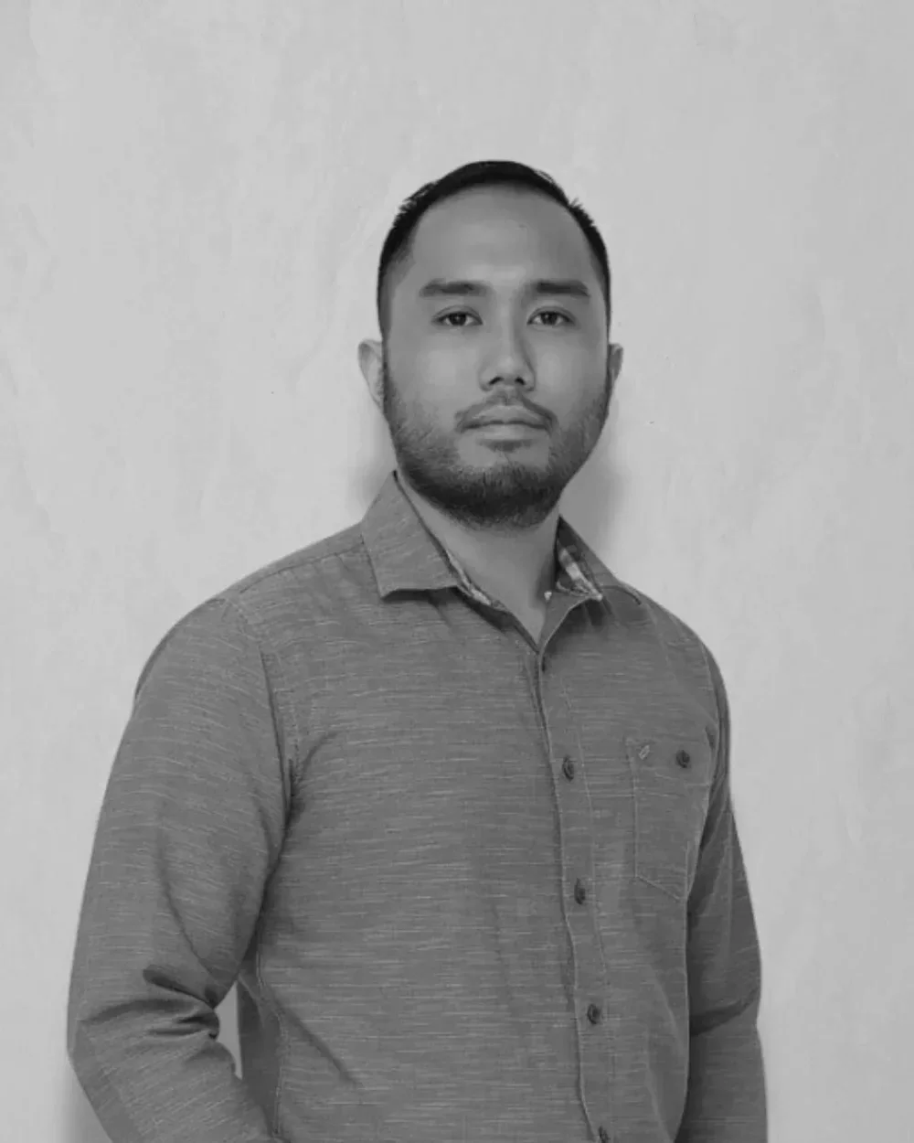 A black and white portrait of a man with short dark hair and a beard, wearing a button-up shirt, standing against a plain light-colored background.