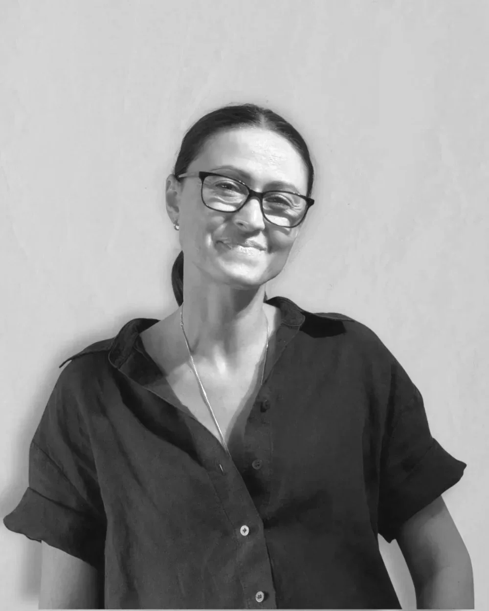 A smiling woman with glasses and dark hair tied back, wearing a dark button-up shirt, standing against a plain wall.