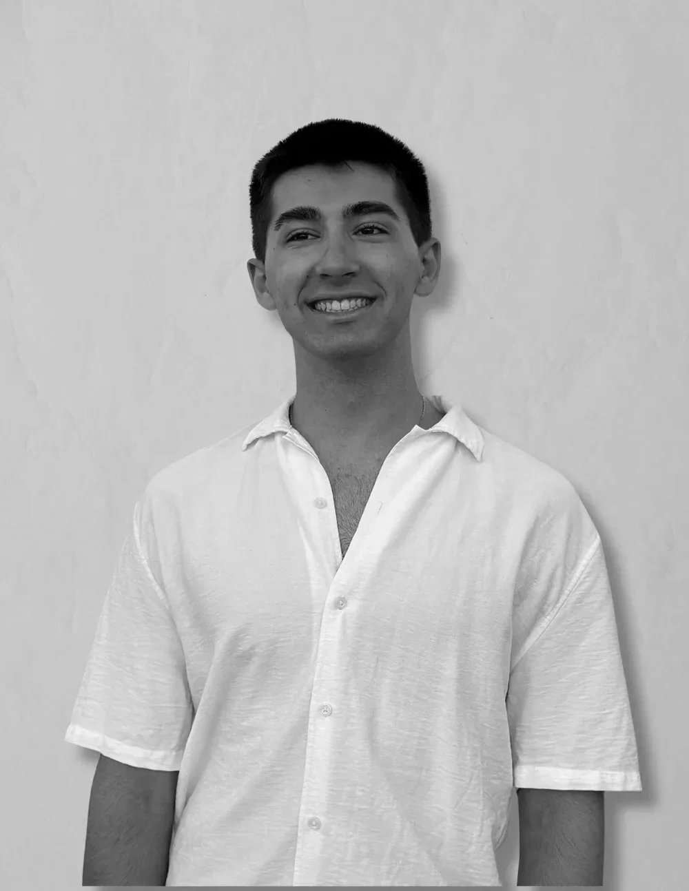 A young man smiling and standing against a plain wall, wearing a white short-sleeved button-up shirt.