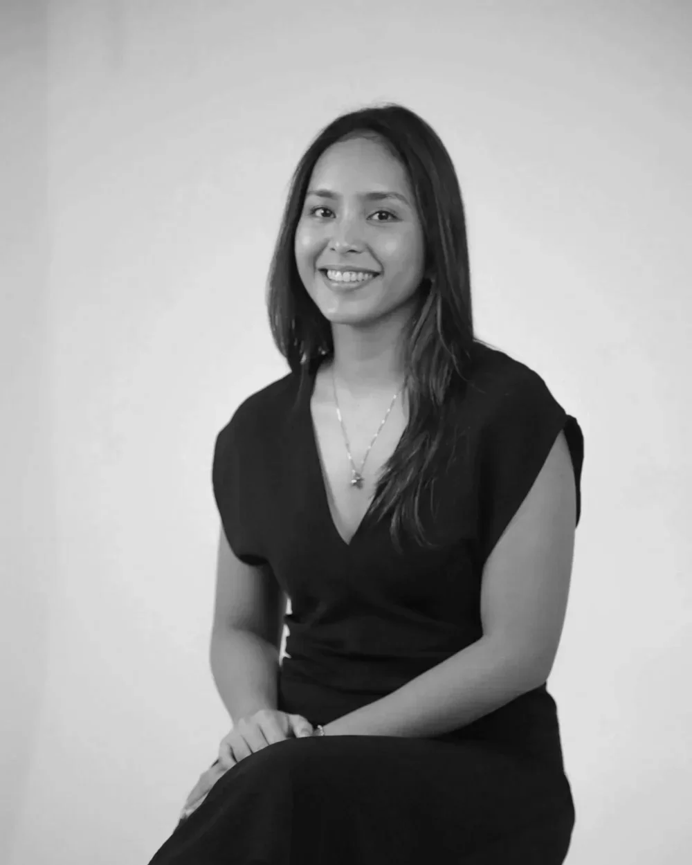 A black and white portrait of a smiling woman with long dark hair, wearing a black top and necklace, sitting on a chair against a plain background.