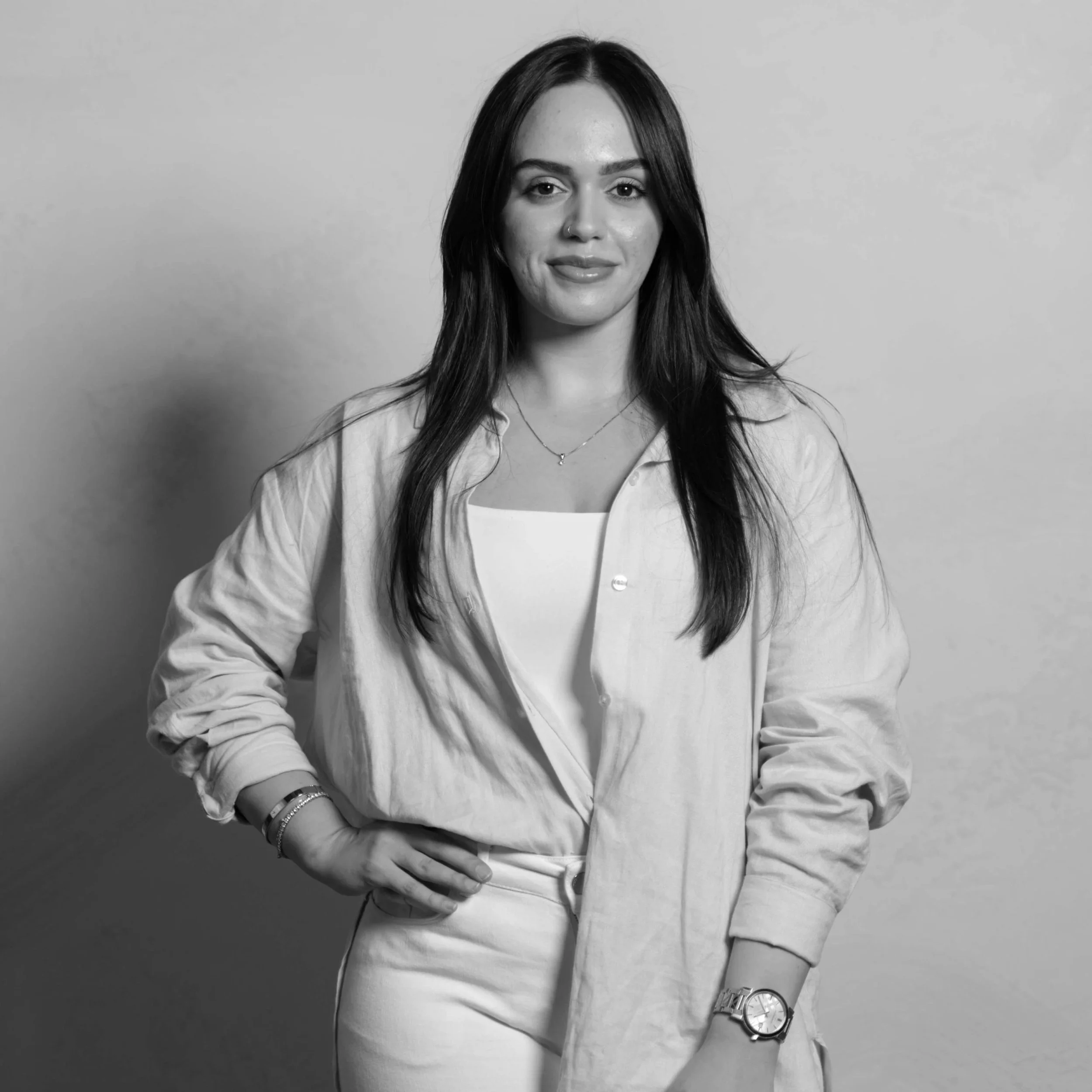 Black and white portrait of a young woman with long dark hair, wearing a light-colored shirt over a white top, accessorized with a watch, bracelets, a necklace, and a nose ring, standing against a plain background.