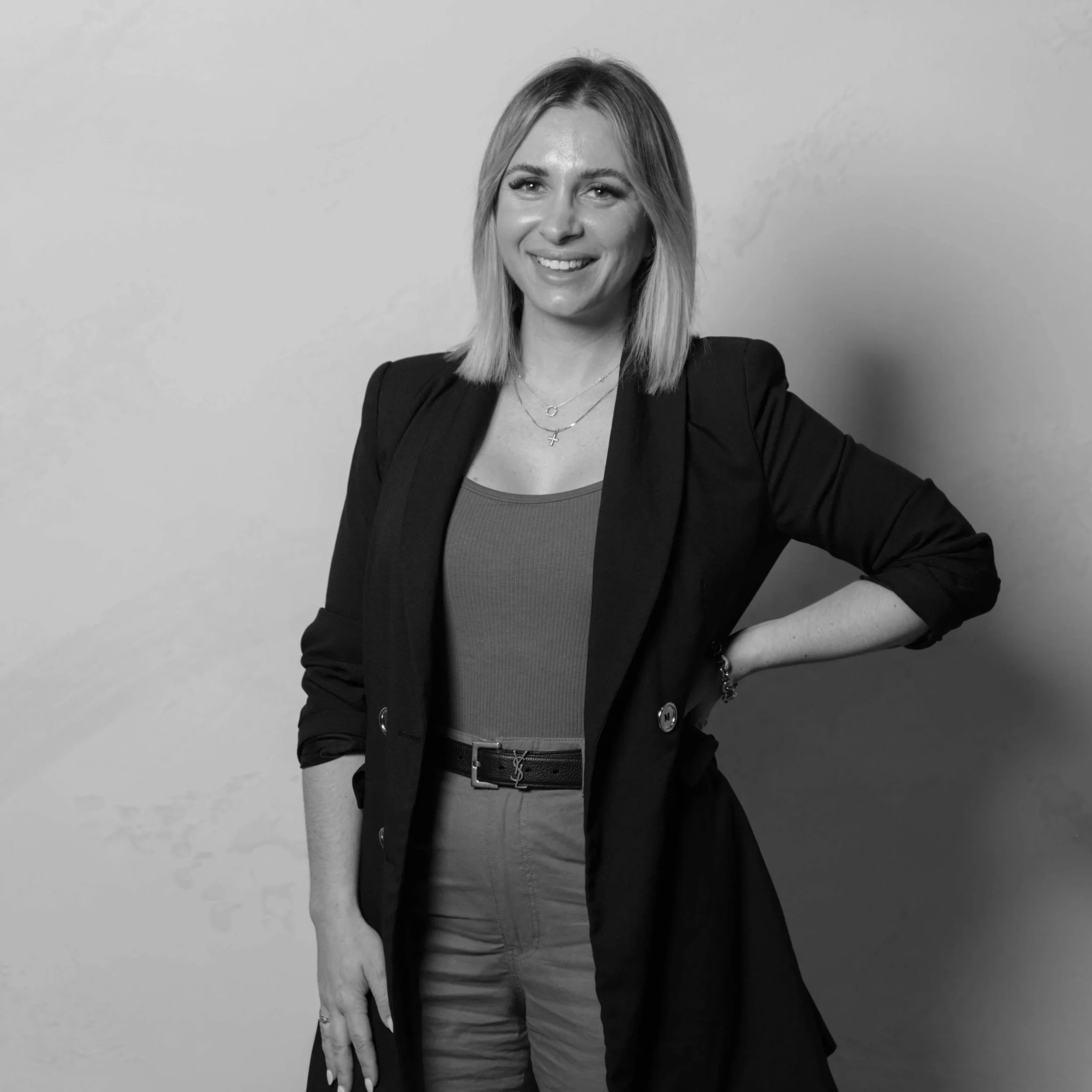 Black and white photo of a woman with shoulder-length hair smiling, wearing a blazer over a top, with a belt, and standing against a plain wall.
