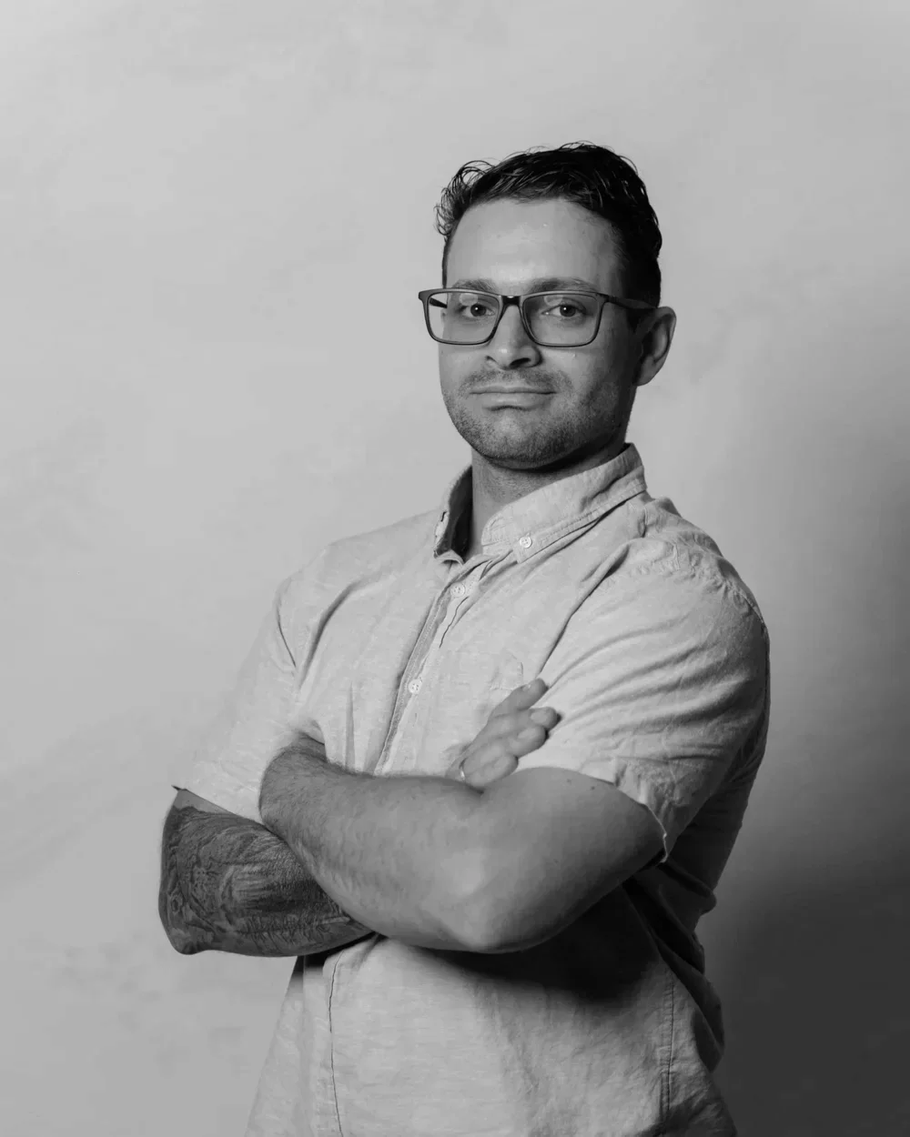Black and white portrait of a man wearing glasses and a button-up shirt, posing with arms crossed against a plain background.