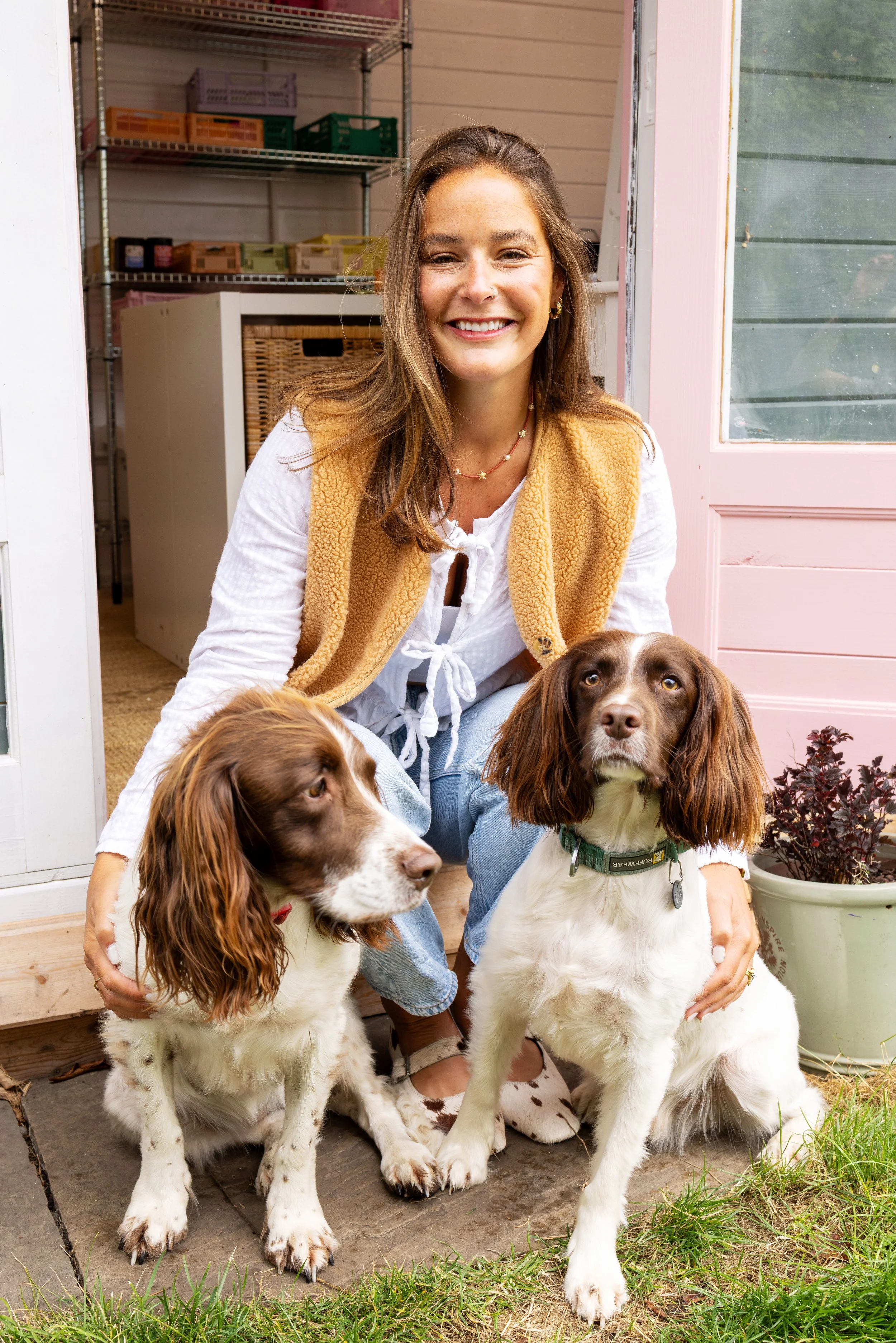 A woman crouches outdoors near a door with pink trim, smiling at the camera. She has brown hair, wears a white long-sleeve shirt with a tie in the front, a tan fleece vest, and light blue jeans. Two spaniel dogs sit in front of her; one looks to the side with a red collar, the other looks up with a green collar. There is a potted plant with dark purple leaves beside them.