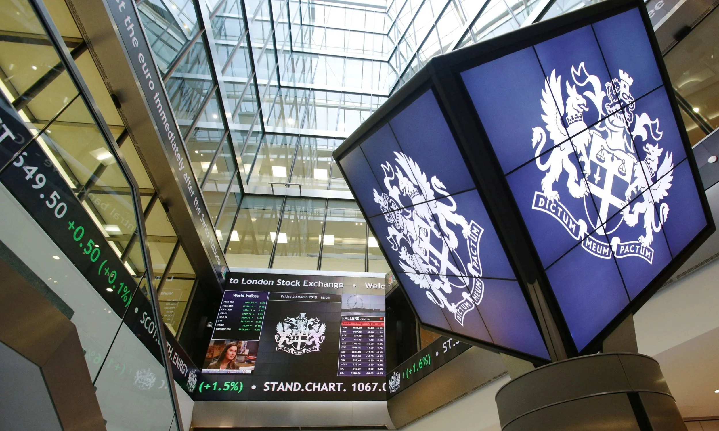 Interior of a financial trading floor or stock exchange with multiple digital screens displaying stock data and logos, including the crest of the London Stock Exchange.