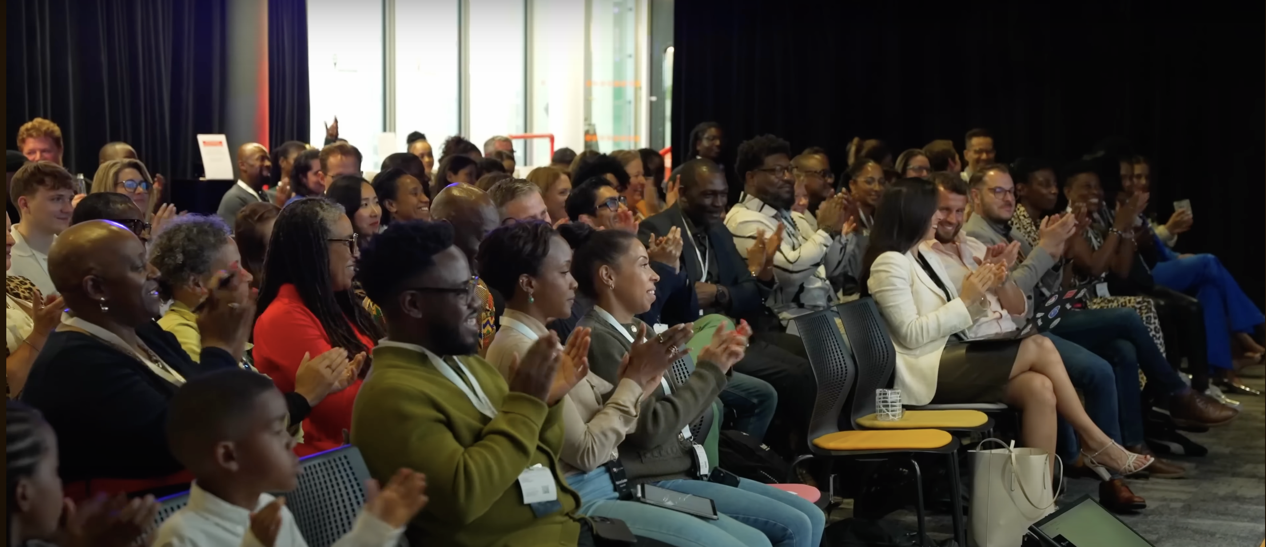 Audience at a conference sitting in rows, clapping and smiling, with some using laptops and phones, in a room with dark curtains and large windows.