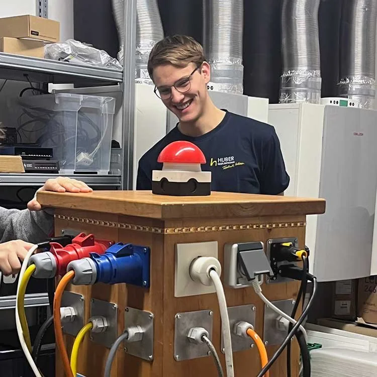 Mann mit Brille lächelt und beobachtet einen roten rosierten Knopf auf einem Holztisch, in einem technischen Raum mit Kabeln und Geräten im Hintergrund.
