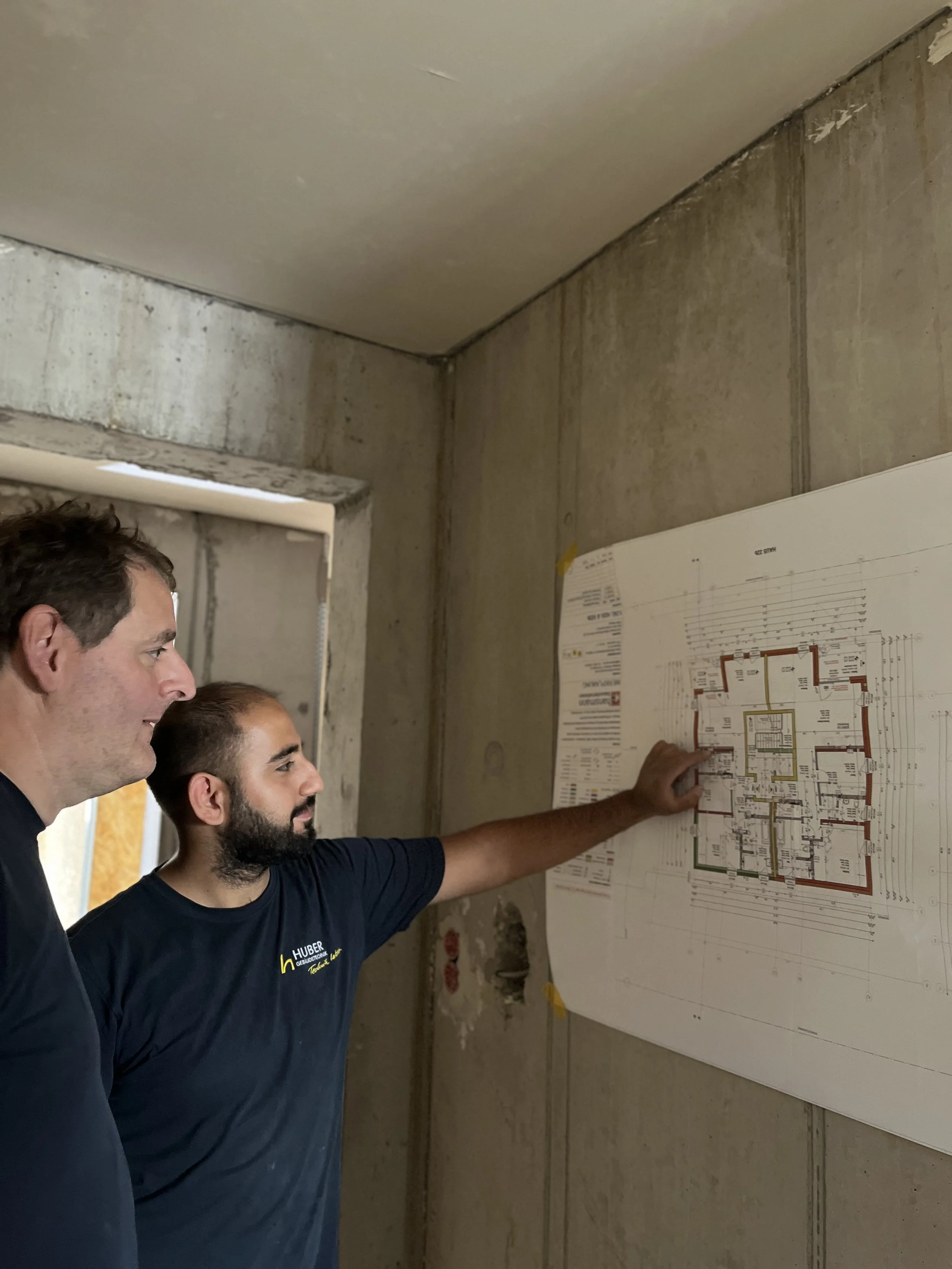 Zwei Männer betrachten eine Grundrisszeichnung an einer rohen Betonwand in einem Bauprojekt. Einer zeigt auf die Skizze, während beide konzentriert zuschauen.
