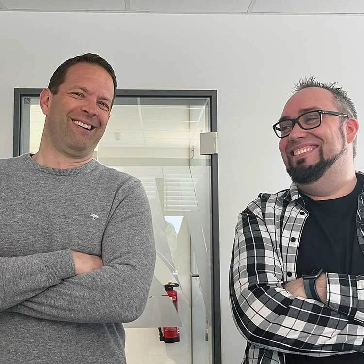 Zwei Männer stehen nebeneinander, lachen und haben die Arme verschränkt in einem Büro.