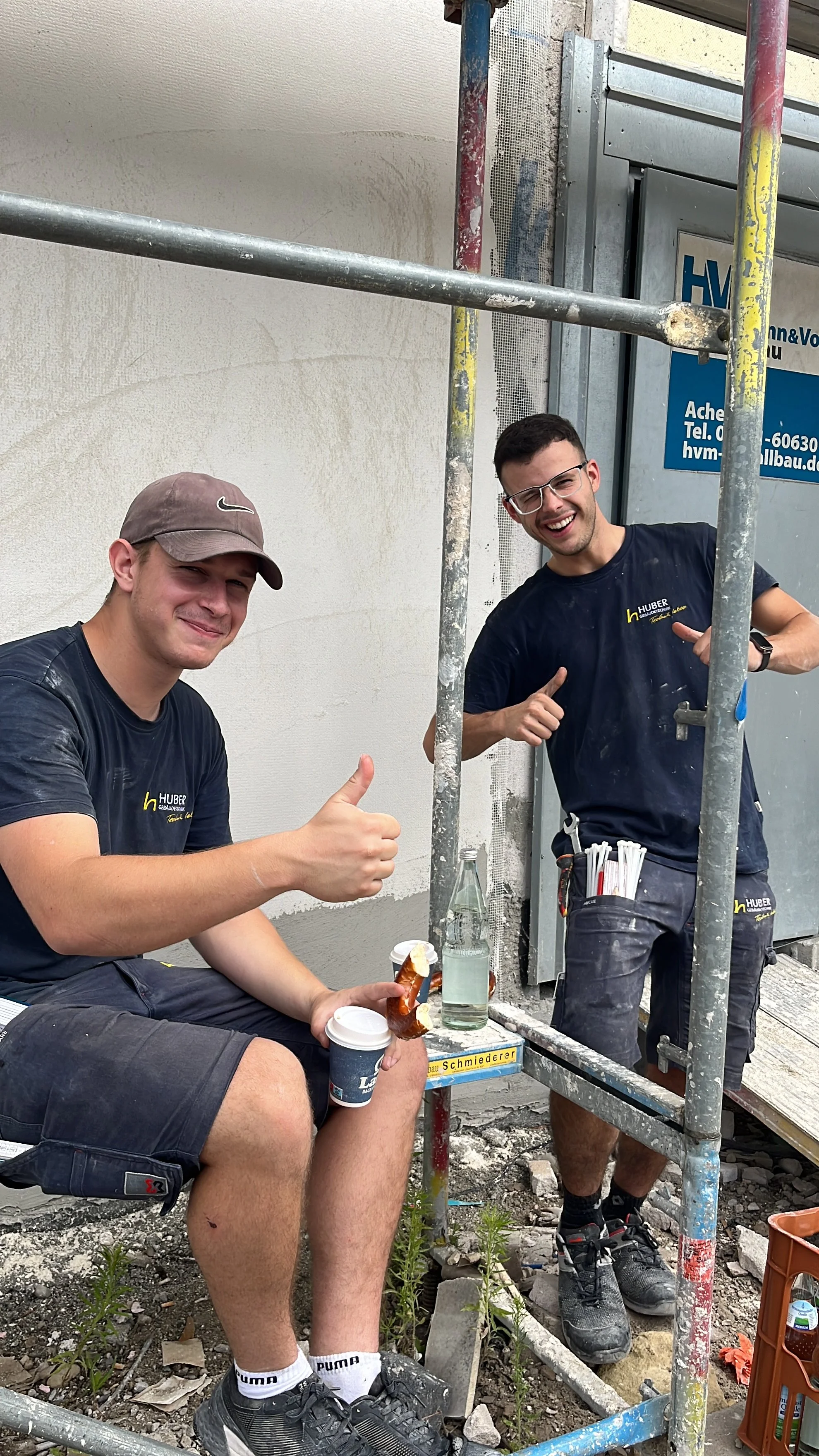 Zwei Männer auf einer Baustelle, lächelnd, einer sitzt mit einem Kaffee und einem Snack, der andere steht in der Gerüst. Beide tragen schwarze T-Shirts mit einem Firmenlogo, lachen und zeigen Daumen hoch.