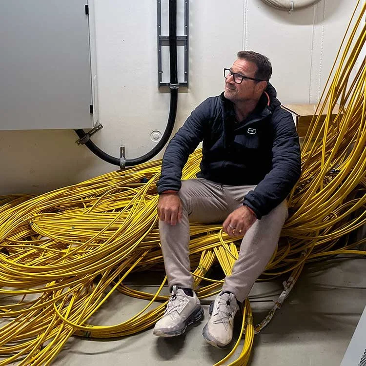 Ein Mann sitzt auf einer Ansammlung von gelben Kabeln in einem technischen Raum.