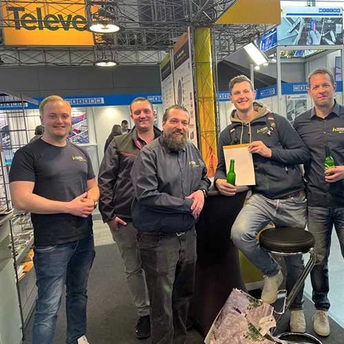 Fünf Männer stehen hinter einem Messetresen bei einer Messe, einige halten Getränke in der Hand. Im Hintergrund sind Ausstellerstände und Leuchtanzeigen zu sehen, darunter ein gelbes "Televes"-Schild.