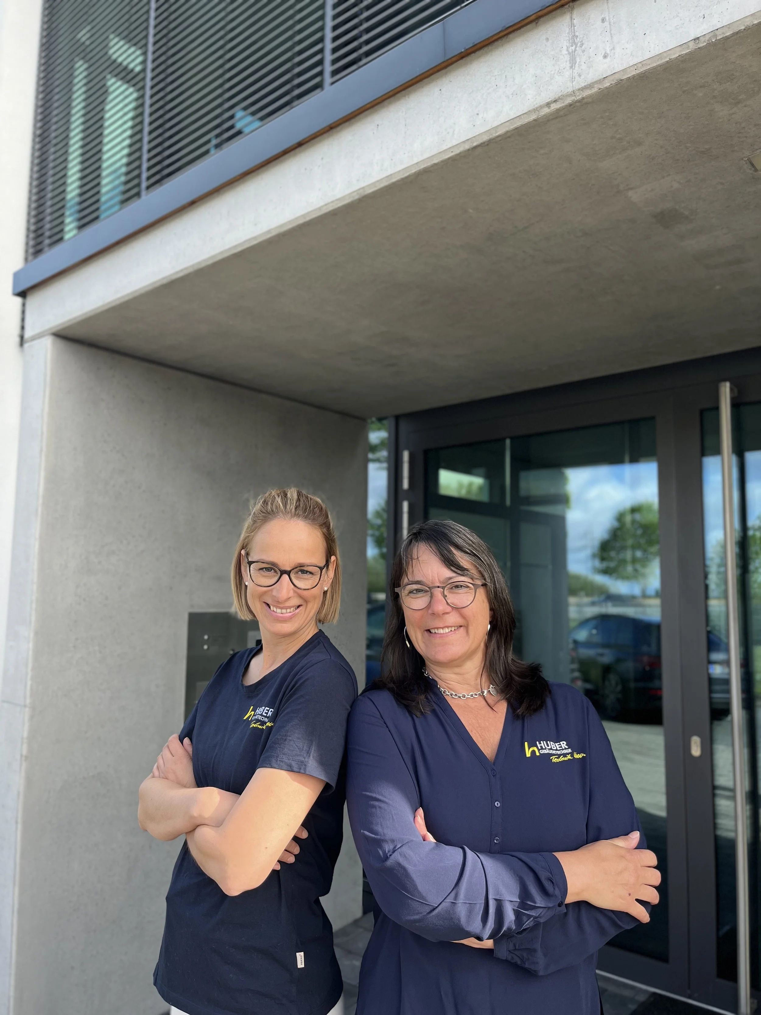 Zwei Frauen stehen vor einem modernen Gebäude mit schwarzen Türen und großen Fenstern, beide tragen dunkelblaue Shirts mit einem Firmenlogo.