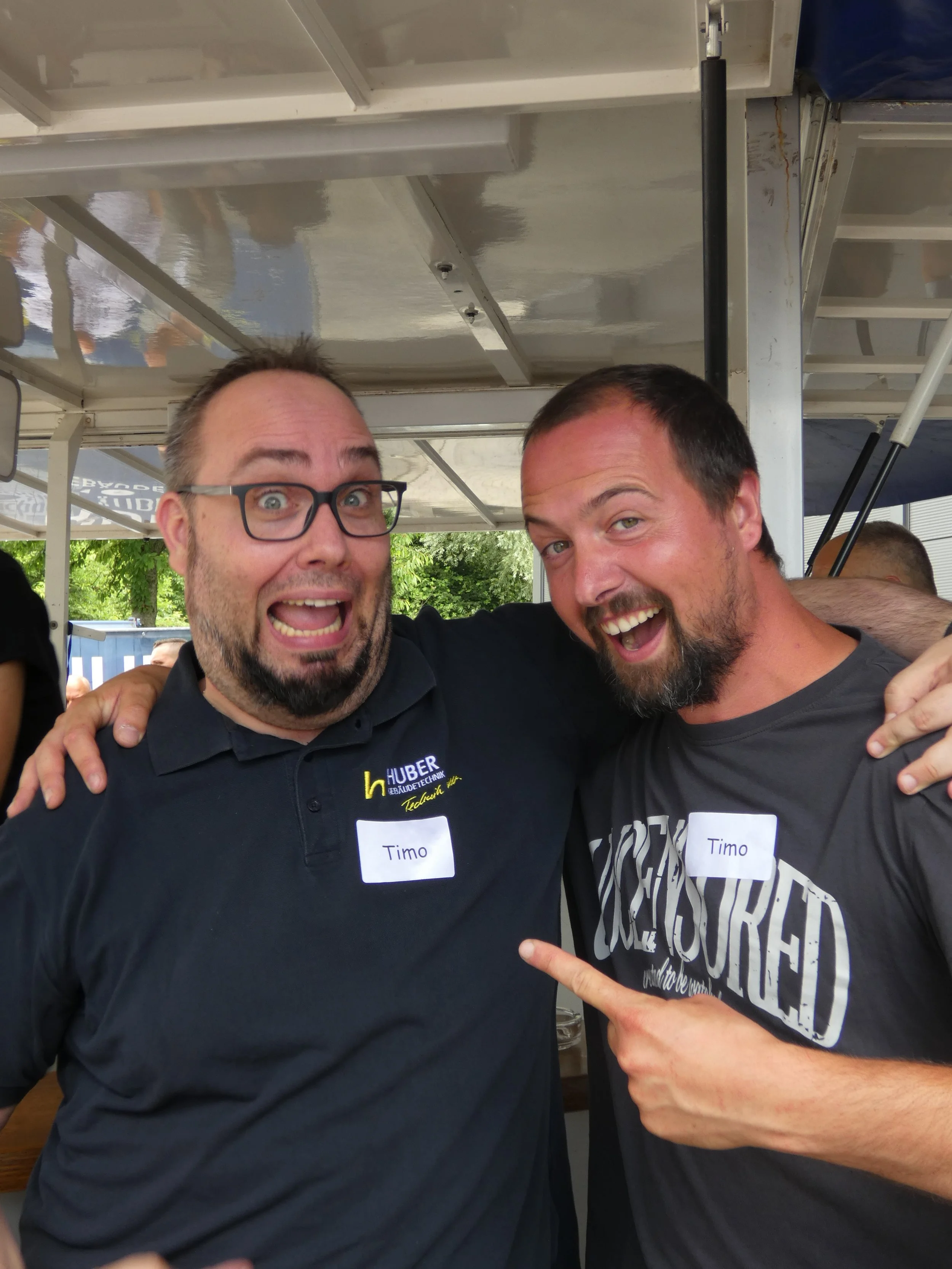 Zwei lachende Männer, einer trägt eine Brille und ein schwarzes T-Shirt mit einem Namensschild 'Timo', der andere trägt ein schwarzes T-Shirt mit Schriftzug. Beide haben die Arme um die Schultern und zeigen nach vorne, sie sind auf einem Fest oder Ev