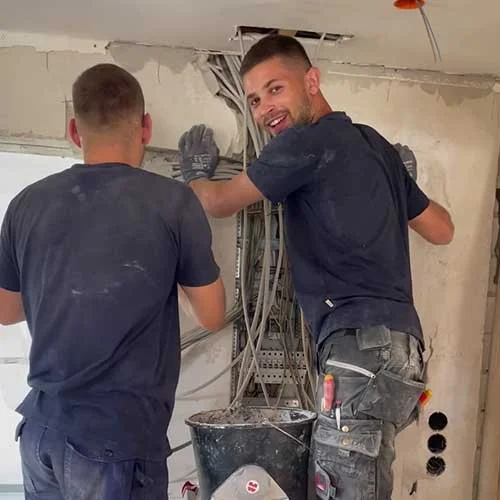 Zwei Männer arbeiten an einem nervösen Elektrikerdienst in einem Raum mit ungeputzter Wand und offenen Kabeln, einer lächelt in die Kamera.