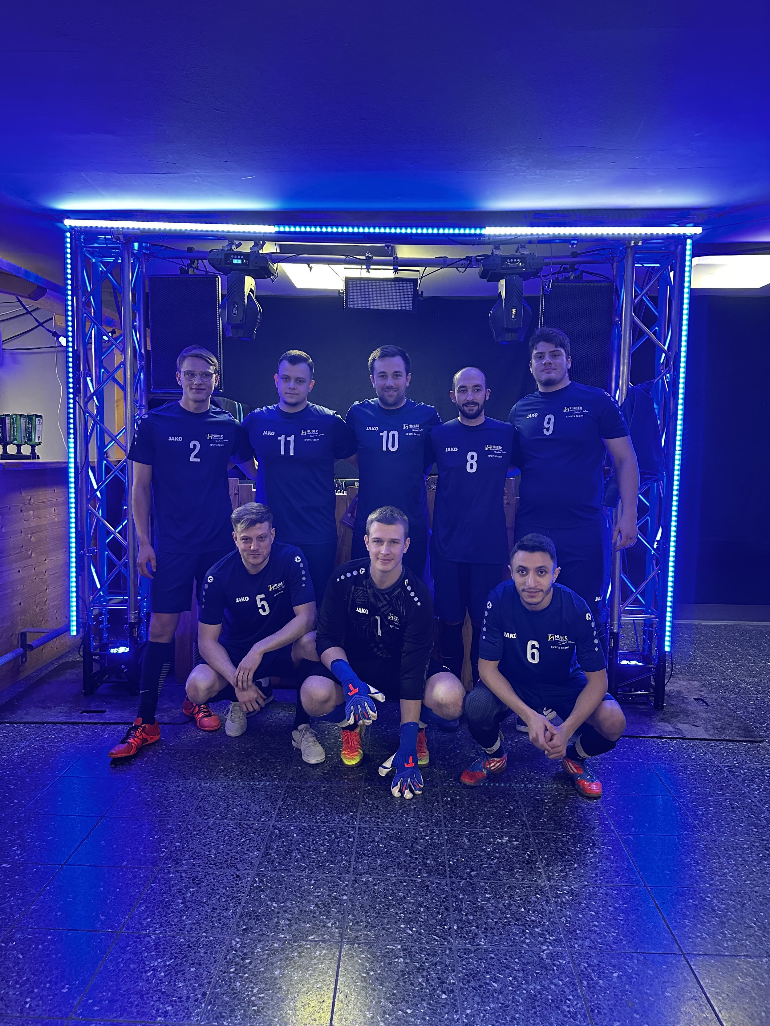 Mannschaftsfoto einer Volleyballmannschaft in blauen Trikots in einem Raum mit blauer Beleuchtung.