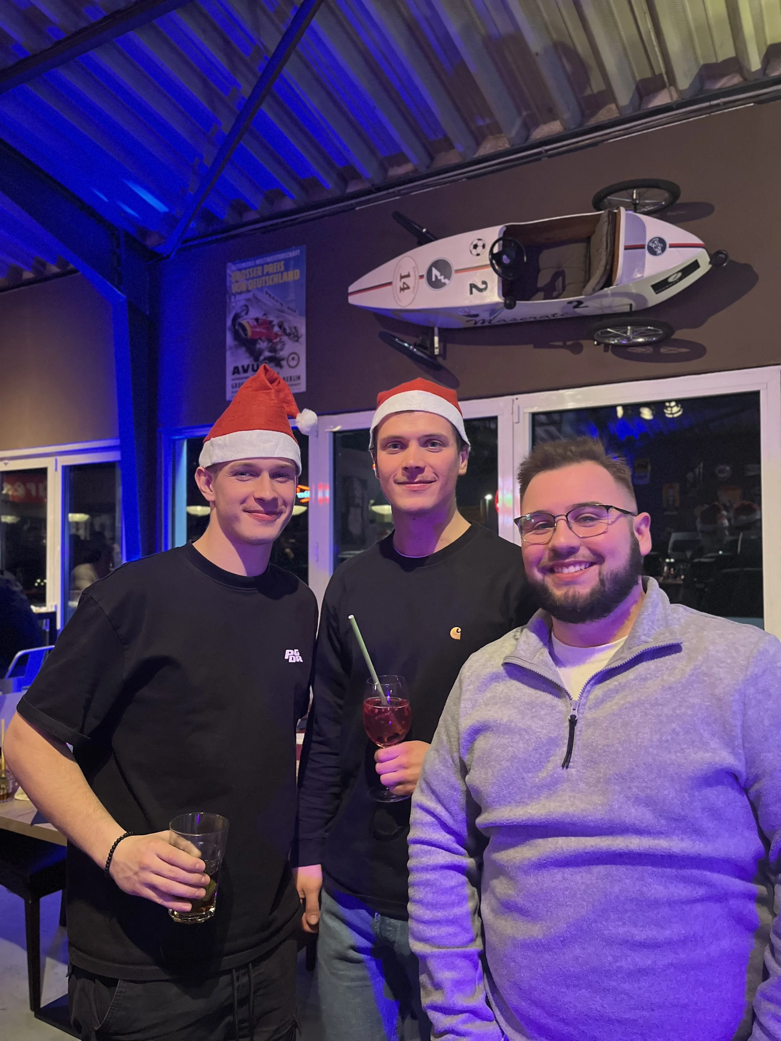 Drei Männer mit Weihnachtsmützen, die in einer Bar oder einem Restaurant feiern. Sie haben Getränke in der Hand und lachen. Es ist eine festliche Atmosphäre mit Weihnachtsdekoration.