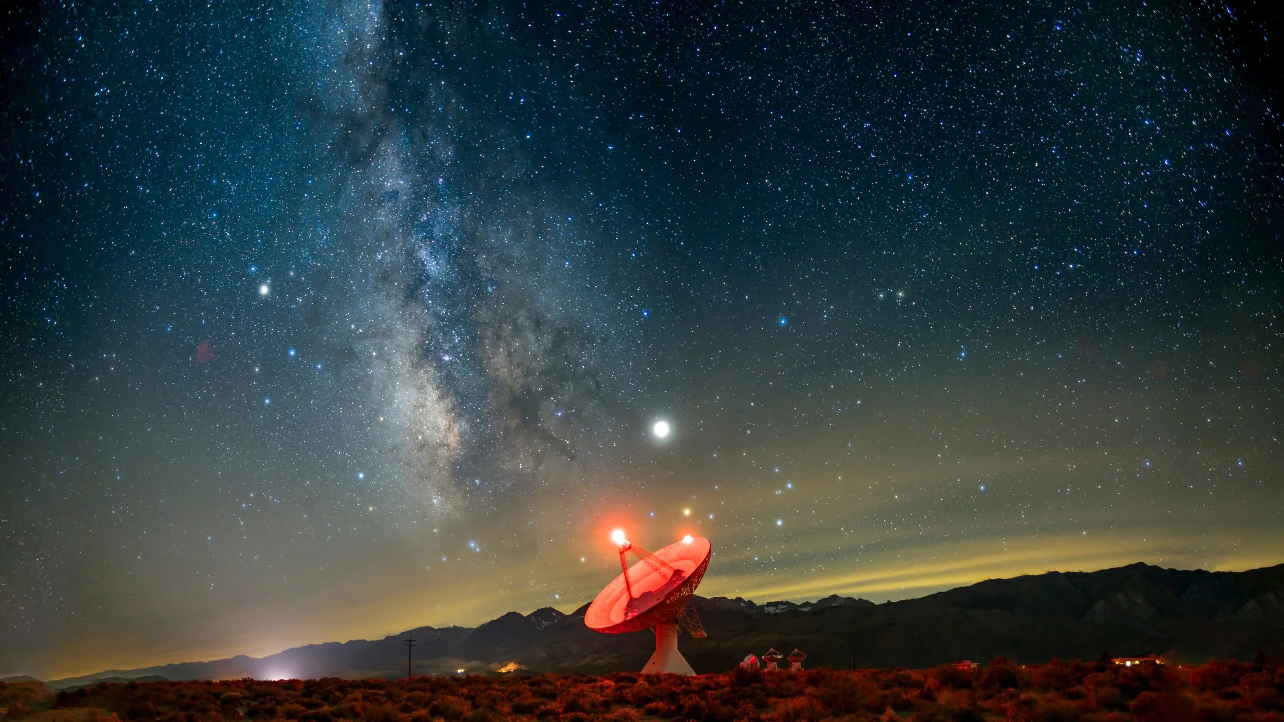 A radio telescope dish pointed towards a star-filled night sky with the Milky Way galaxy visible above mountains.