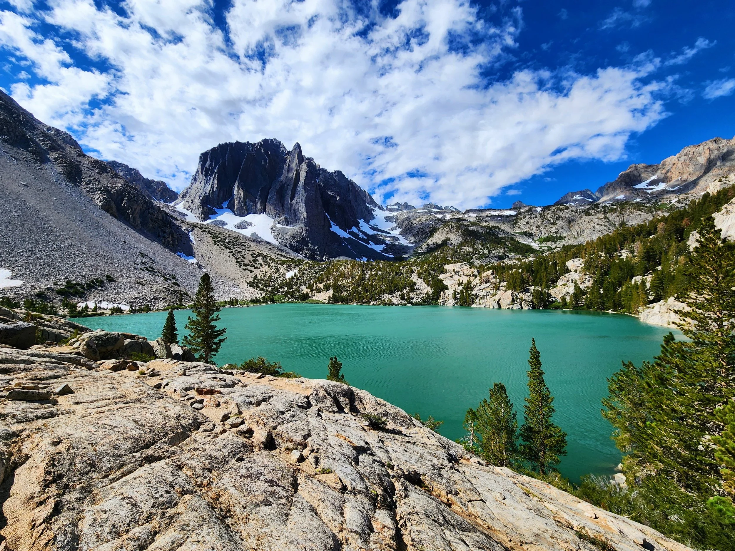 A scenic landscape featuring a turquoise mountain lake surrounded by rocky terrain and green trees, with snow-capped mountains and a partly cloudy blue sky in the background.